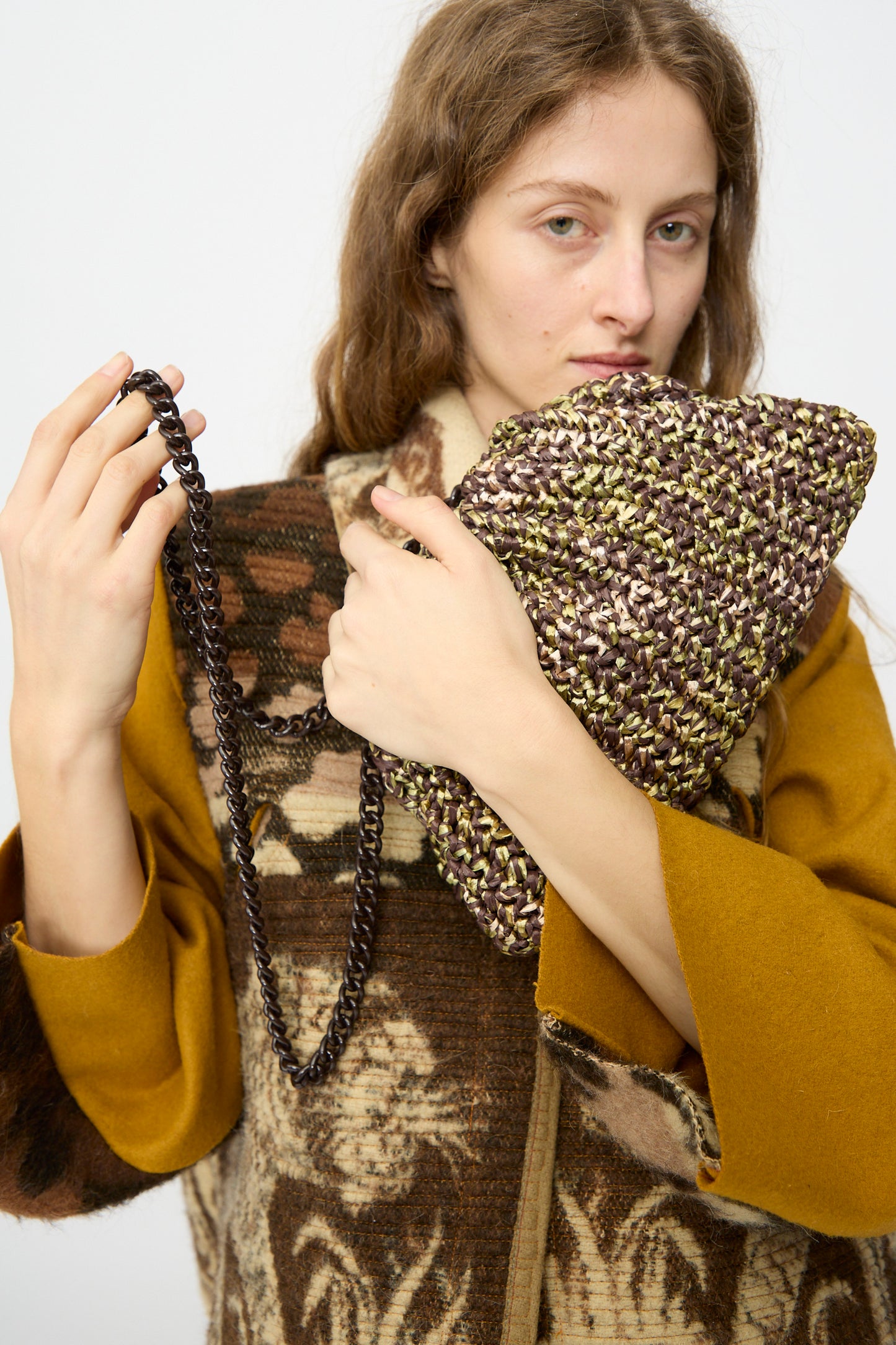 A person with long brown hair holds the Maria La Rosa Earth Game Crochet Bag in Natural, featuring a chain strap, while wearing a brown and yellow patterned coat against a plain background.