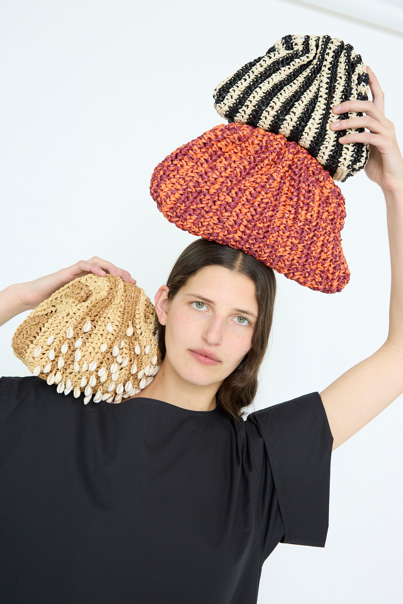 A person holds three Maria La Rosa Shell Mini Game Bags in various styles—beaded natural, red-orange, and black-and-white striped—against a plain backdrop.