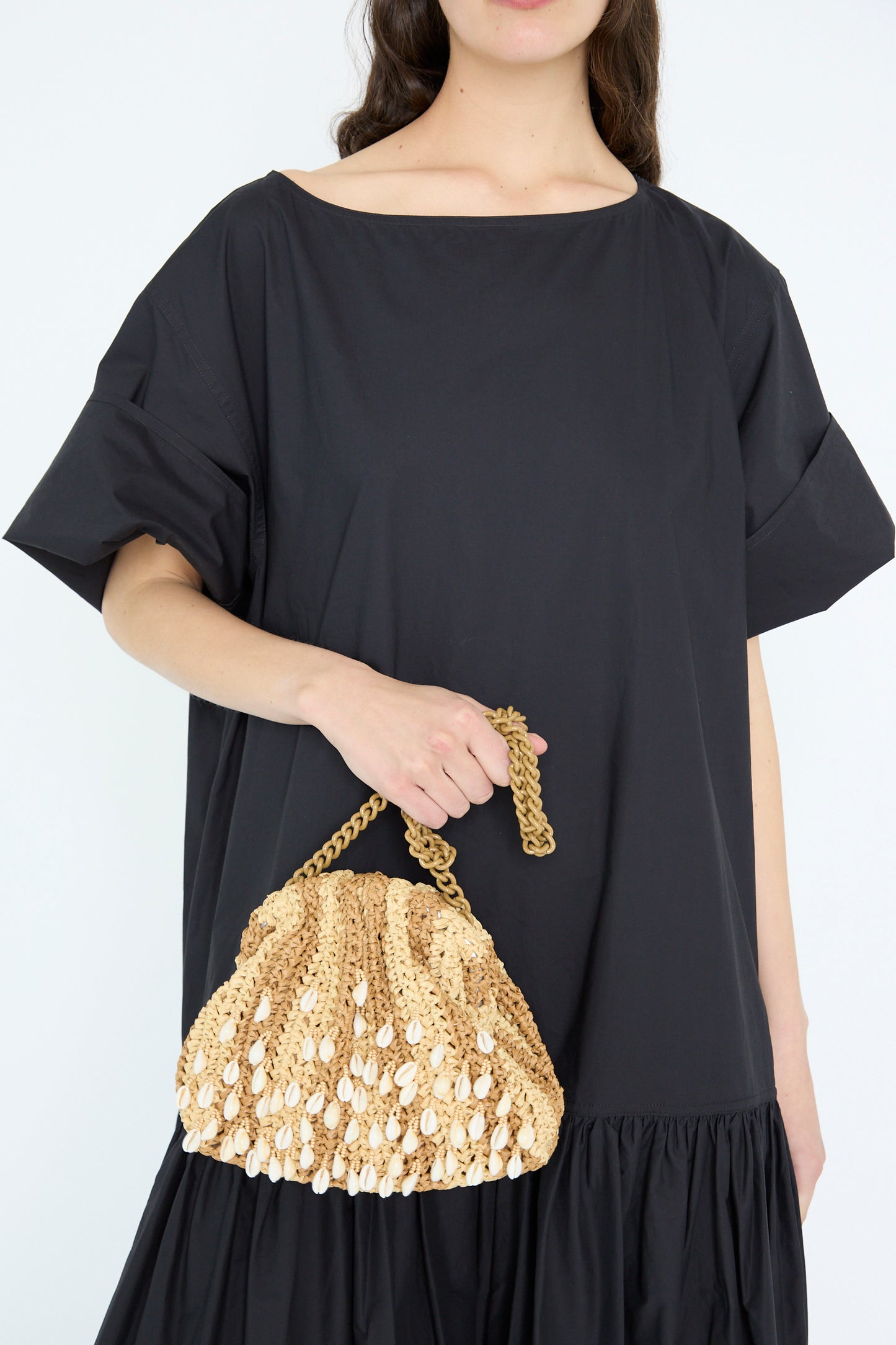 A person in a black dress holds the Shell Mini Game Bag by Maria La Rosa, a beige woven bag with shell details and a chain shoulder strap.