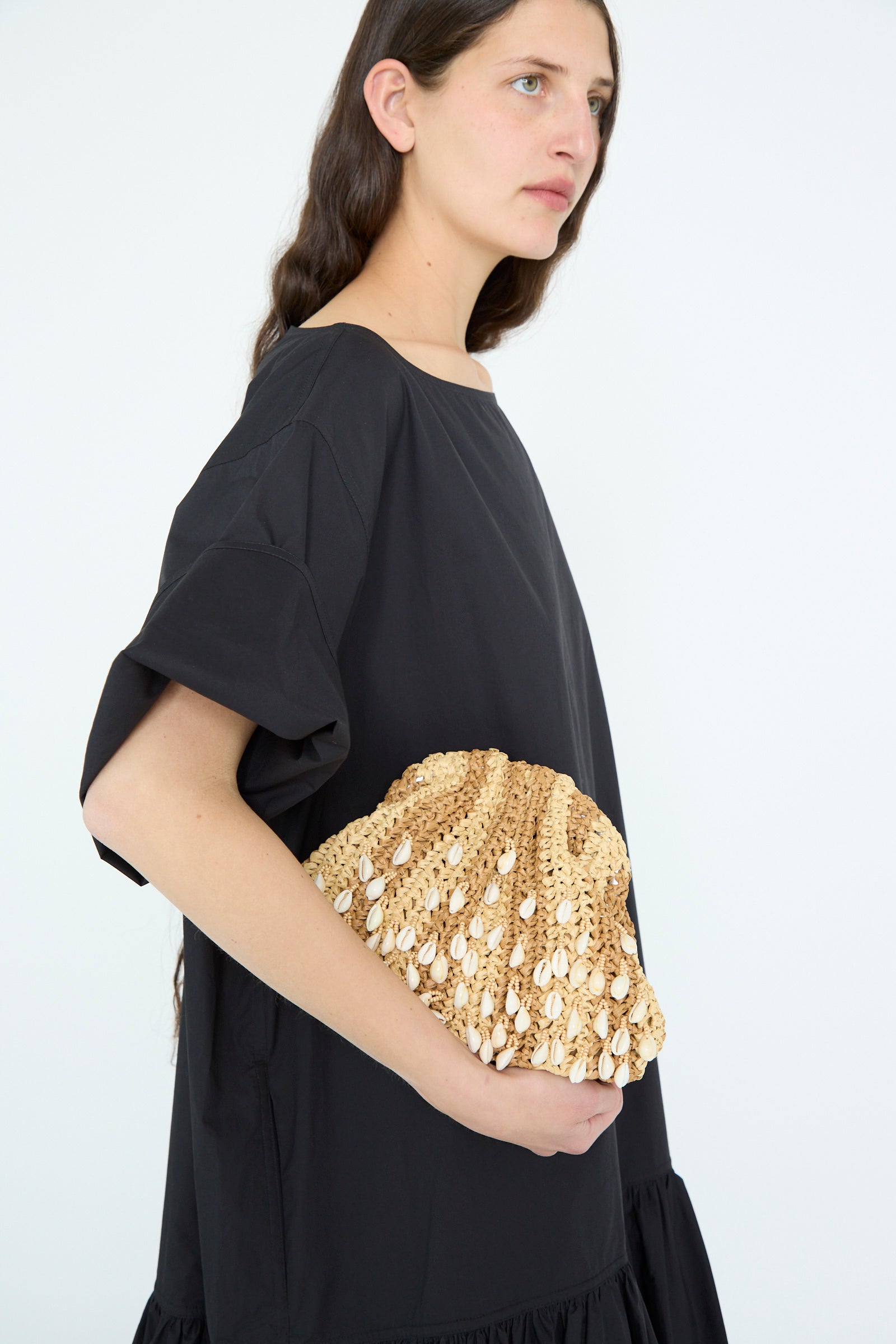 A person in a black dress carries the Shell Mini Game Bag in Natural by Maria La Rosa, featuring a woven straw design with shells and an interior pocket.