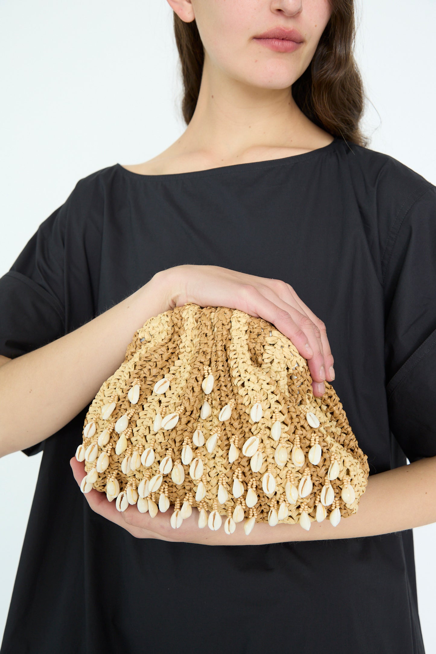 A woman holds the Shell Mini Game Bag in Natural by Maria La Rosa, a woven beige clutch adorned with small shells and featuring an interior pocket, complementing her black top against a plain background.