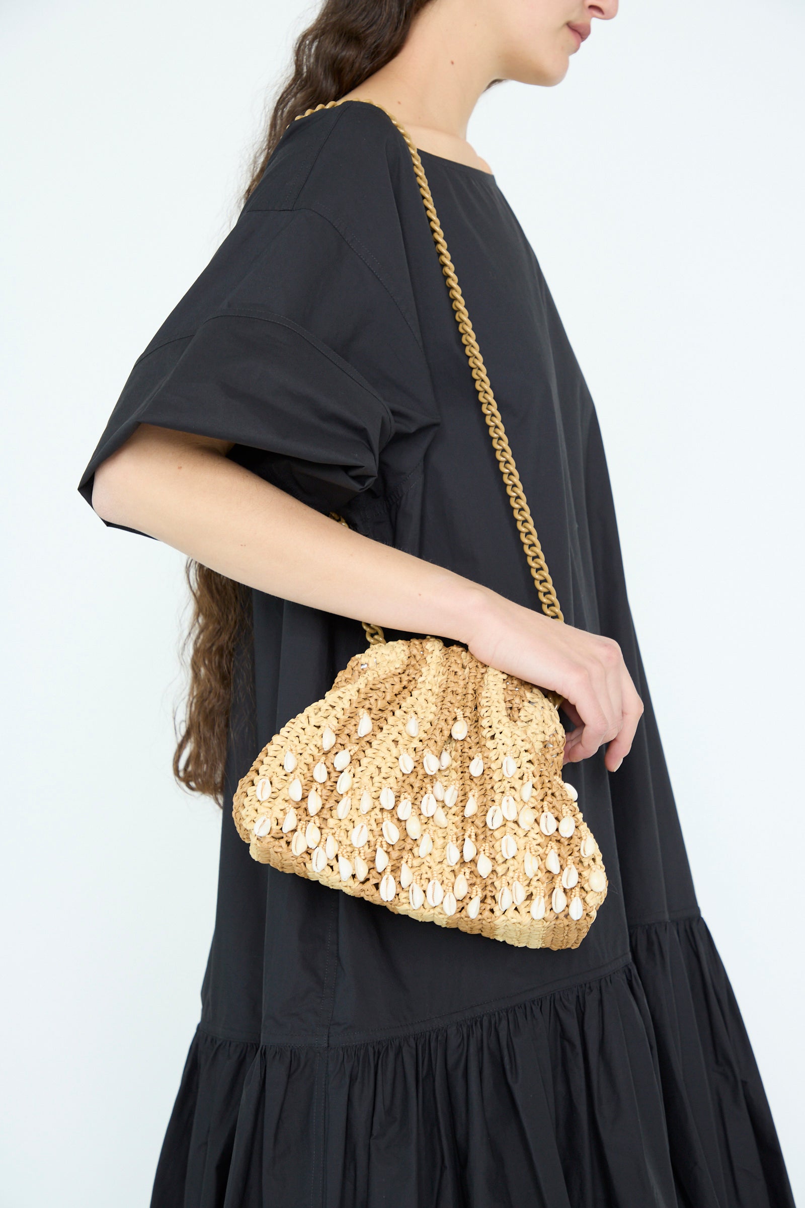 A person in a black dress holds Maria La Rosa's Shell Mini Game Bag in Natural, featuring a scalloped edge with decorative white beads and a chain shoulder strap.