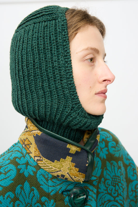 A person in profile wears the Wool Balaclava Lina in Emerald by Maria La Rosa, paired with a patterned scarf featuring yellow and navy accents and a textured blue-green floral coat—showcasing the allure of handmade balaclava style.