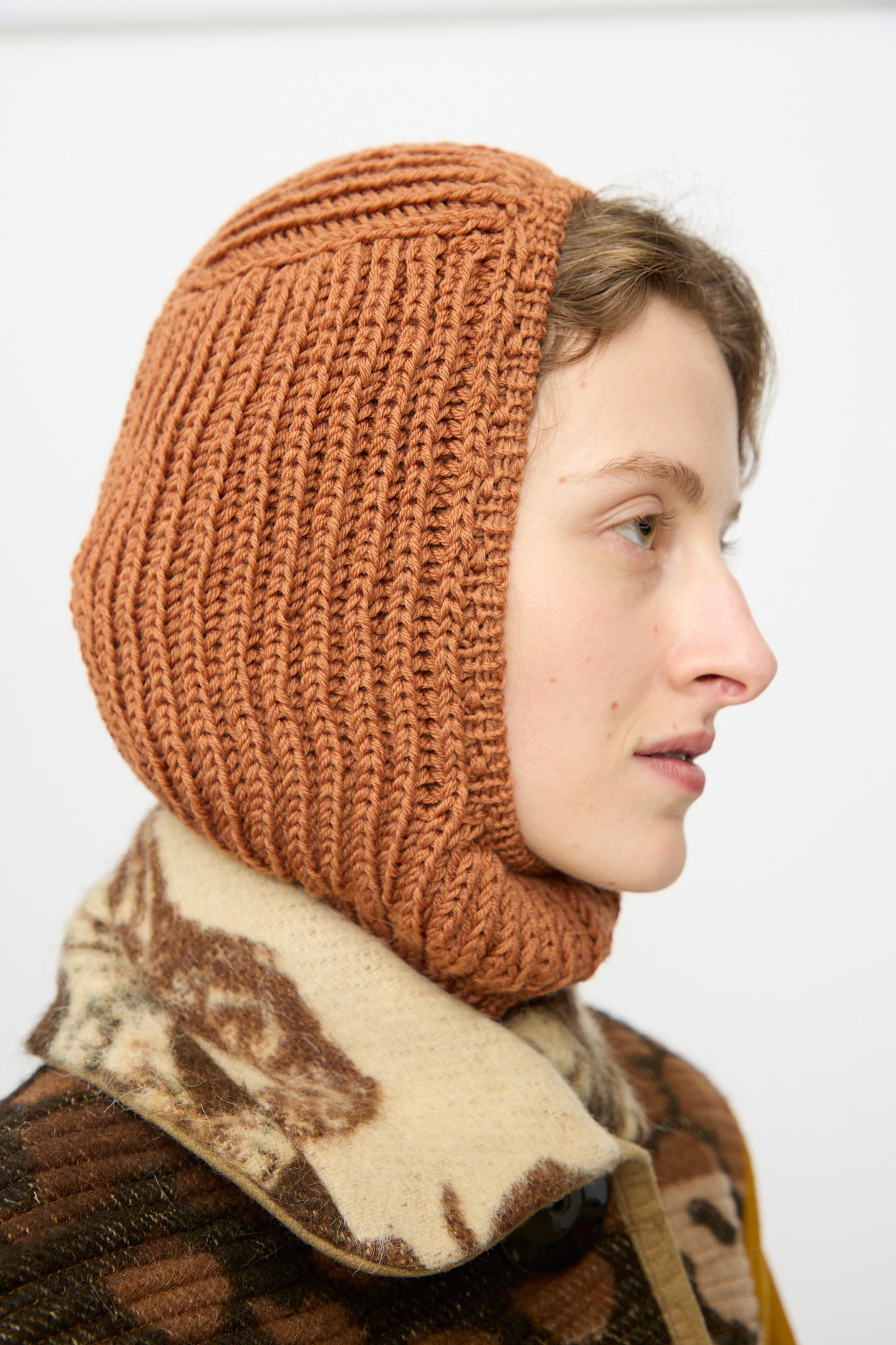 A person in profile wears the Maria La Rosa Wool Balaclava Lina in Rust and a patterned coat, set against a plain background.