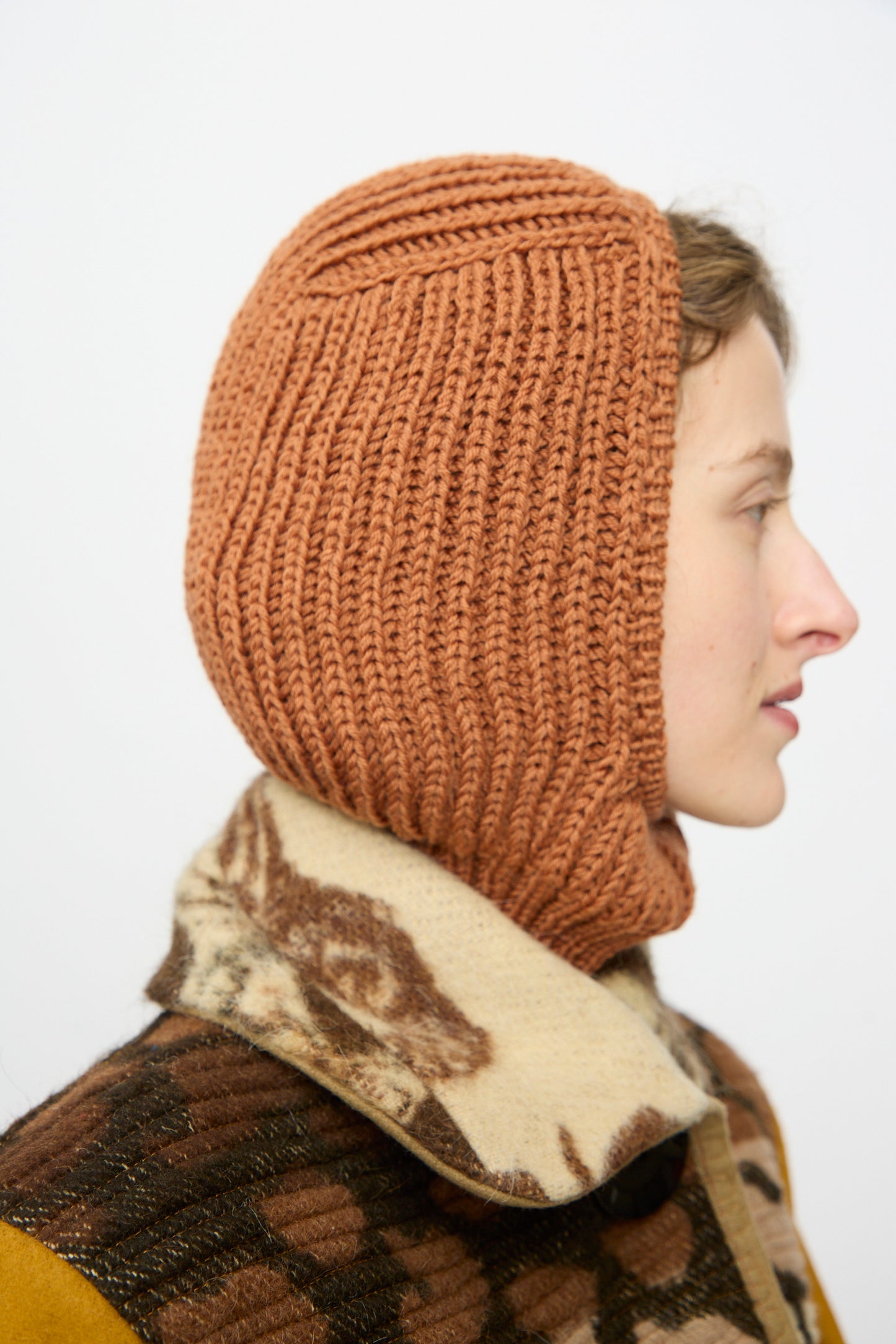 A person in profile wears the Wool Balaclava Lina in Rust by Maria La Rosa and a patterned fleece jacket with a high collar.
