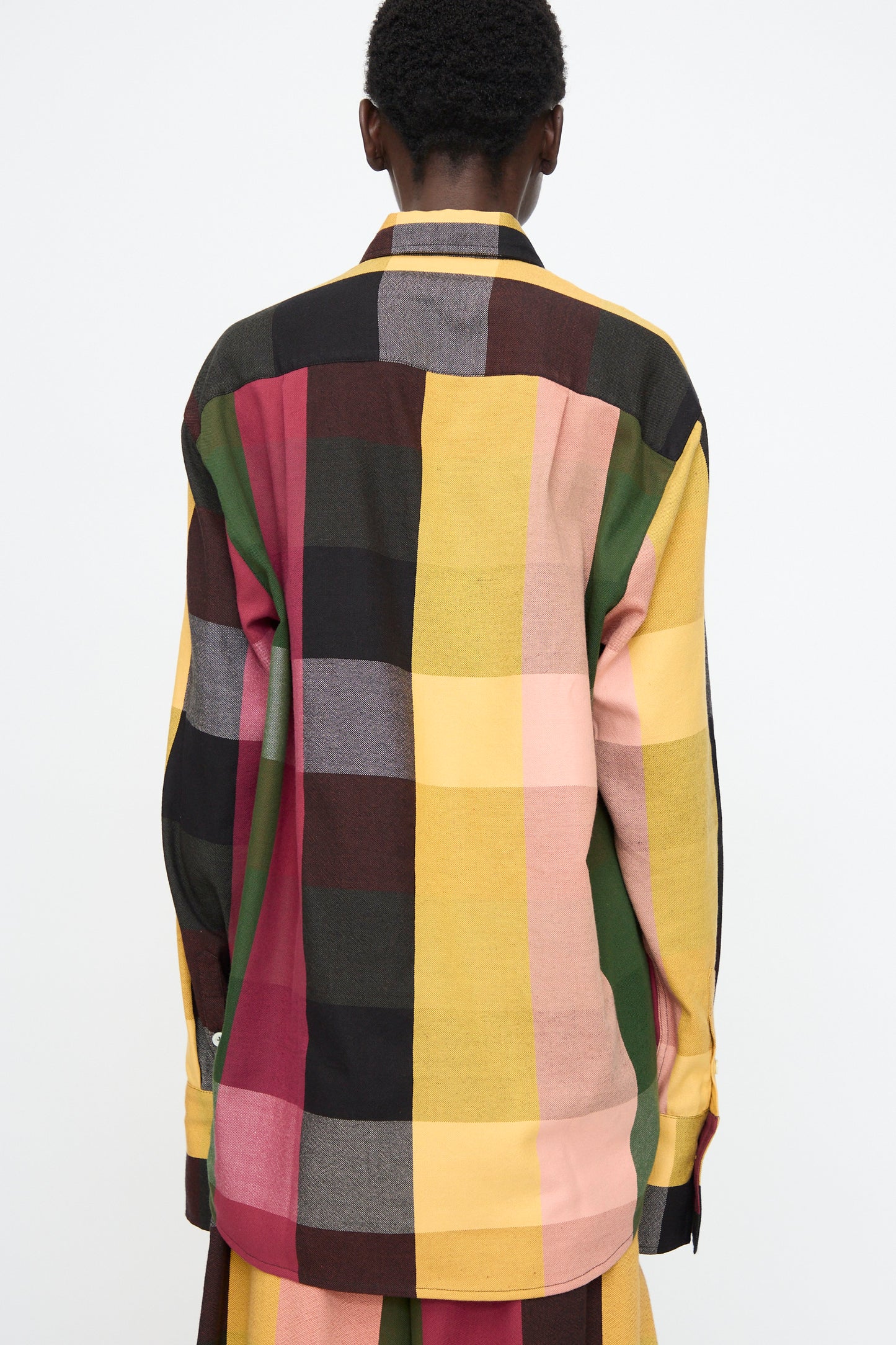 A person stands with their back to the camera wearing the Marrakshi Life Button Down Shirt in Plaid Multi, a relaxed fit long-sleeve shirt with a bold yellow, green, black, red, and beige plaid pattern.