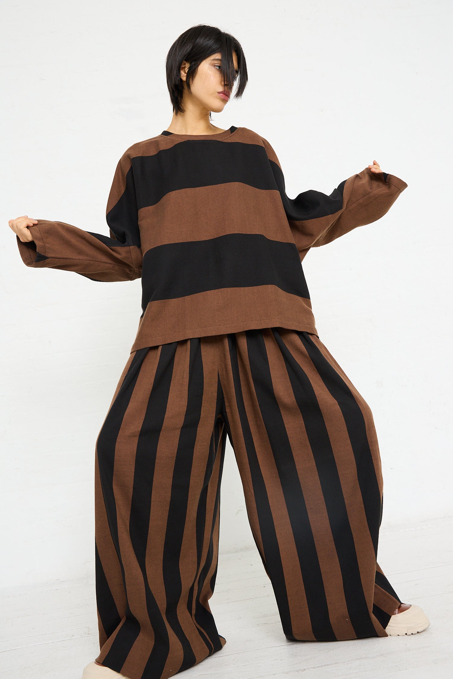 A woman stands against a white background, posing in a loose-fitting, black and brown striped ensemble featuring the Drawstring Pleated Pant in Black and Brown Stripe by Marrakshi Life.