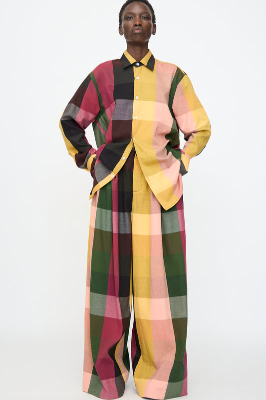 Person standing against a plain background, wearing an oversized shirt and Marrakshi Life's High Waisted Pleated Trouser in Plaid Multi—a wide-leg style featuring a large multicolor plaid cotton pattern in pink, yellow, green, brown, and black.
