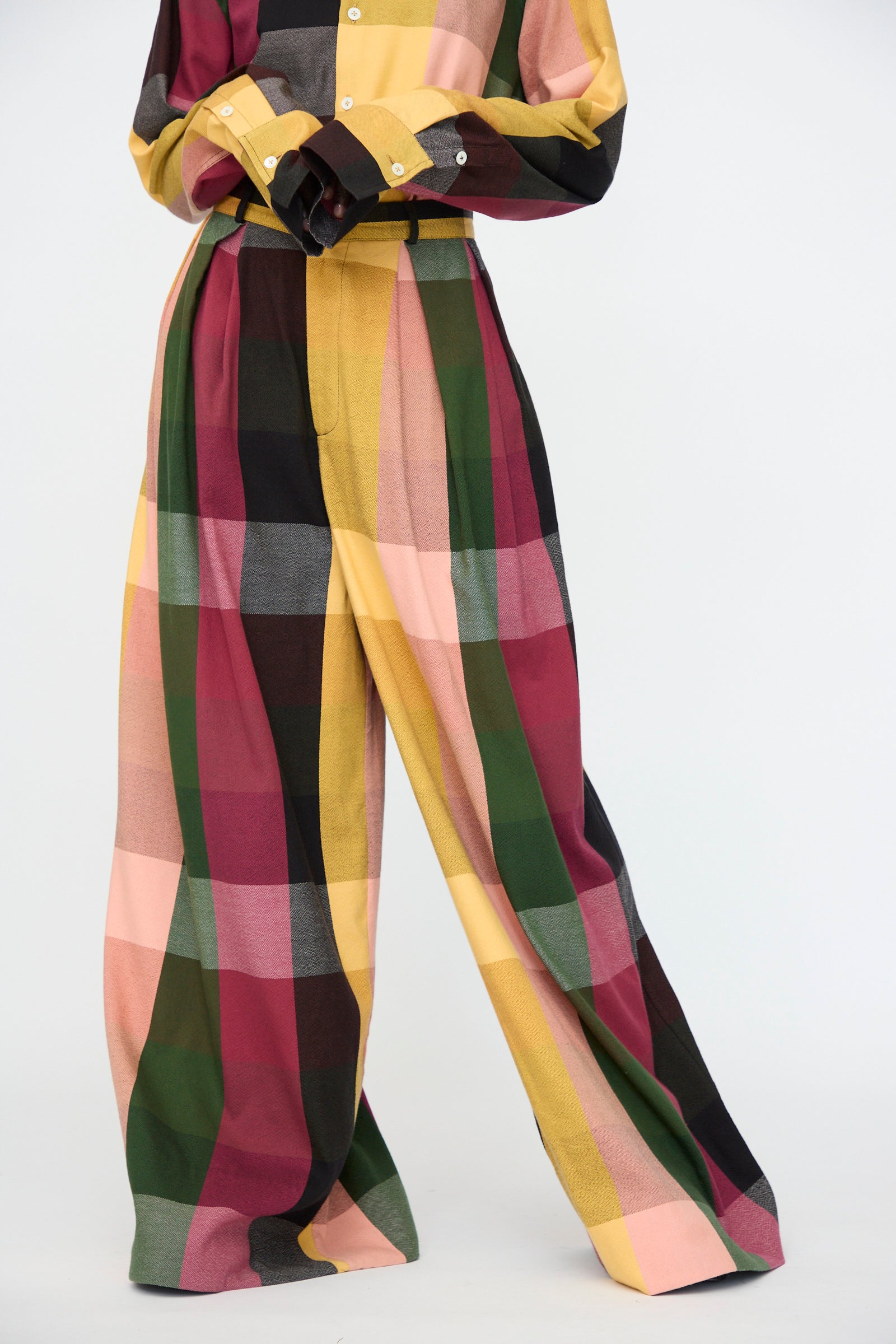A person wears the Marrakshi Life High Waisted Pleated Trouser in Plaid Multi—featuring a bold yellow, green, burgundy, pink, and black checked cotton pattern—against a plain background.