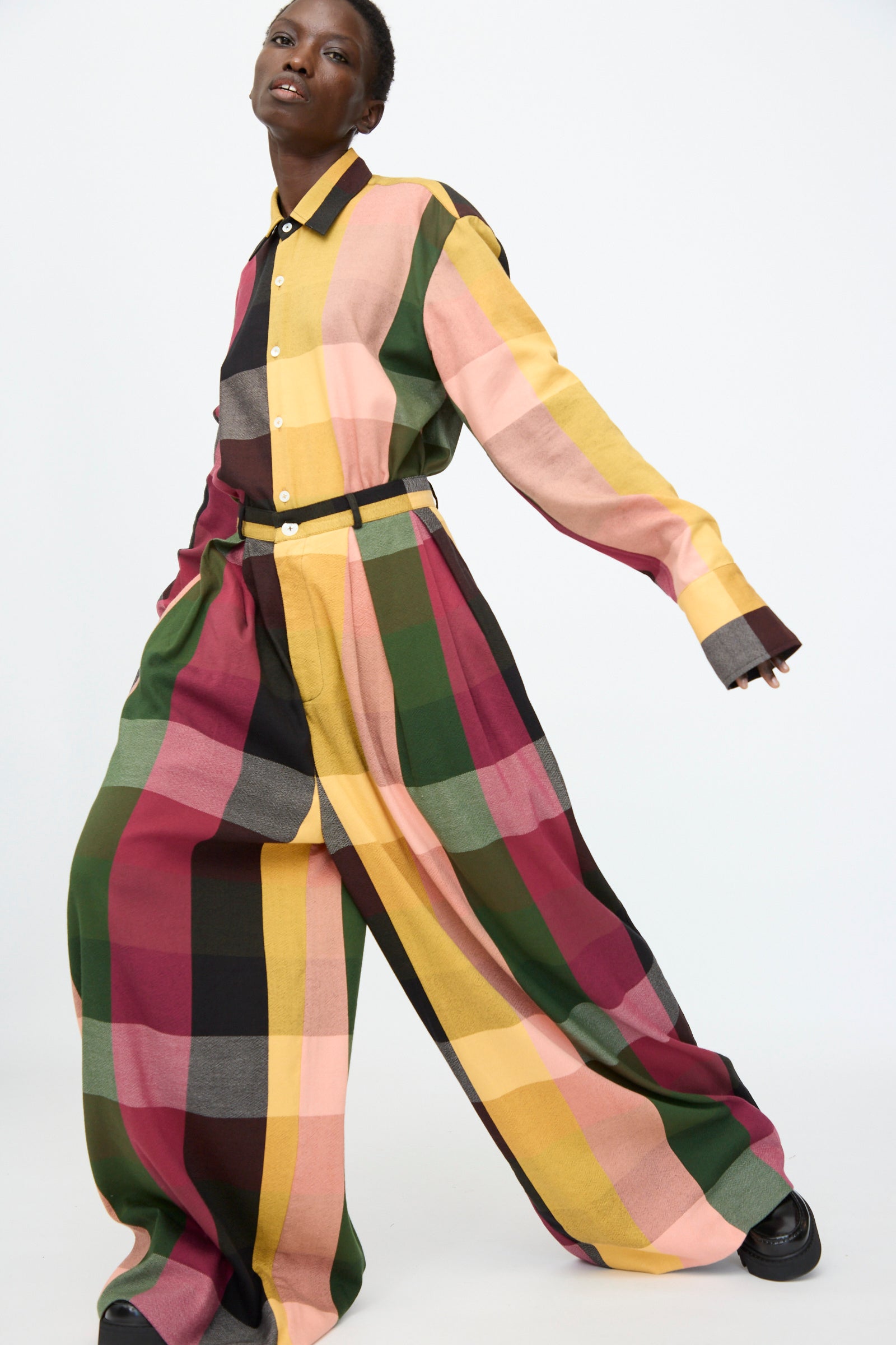 A model wears the Marrakshi Life High Waisted Pleated Trouser in Plaid Multi with a loose, multicolored plaid cotton shirt, posing against a plain white background.