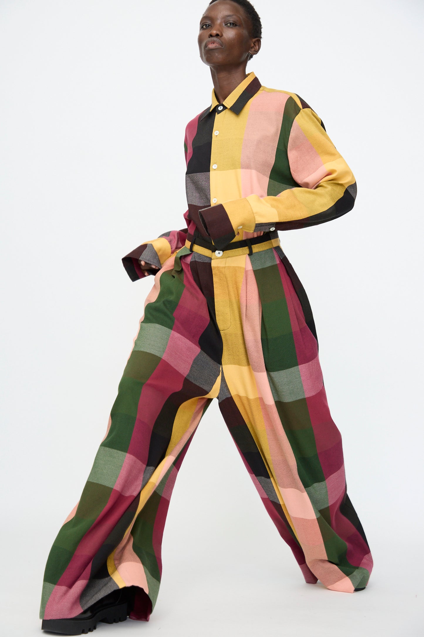 A person wearing Marrakshi Life's High Waisted Pleated Trouser in Plaid Multi, paired with a matching long-sleeve shirt, walks against a plain white background.