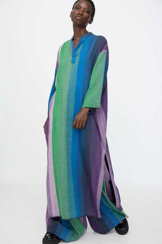 A person wears the Marrakshi Life Nero Collar Caftan in Stripe Multi— a loose, long cotton caftan with green, blue, and purple stripes—standing against a plain white background.