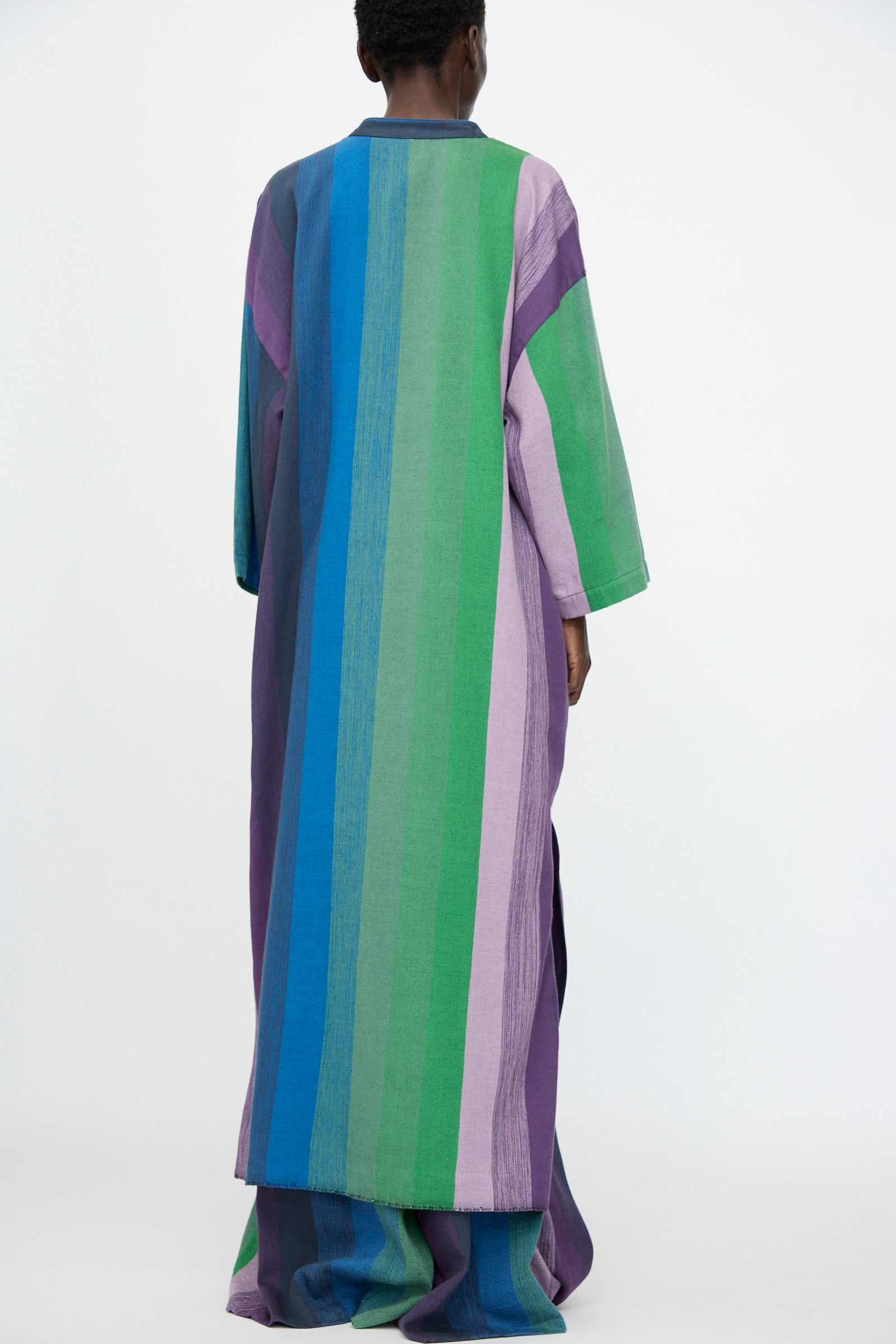 A person stands with their back to the camera, wearing the Marrakshi Life Nero Collar Caftan in Stripe Multi—an oversized caftan with vertical blue, green, and purple stripes.