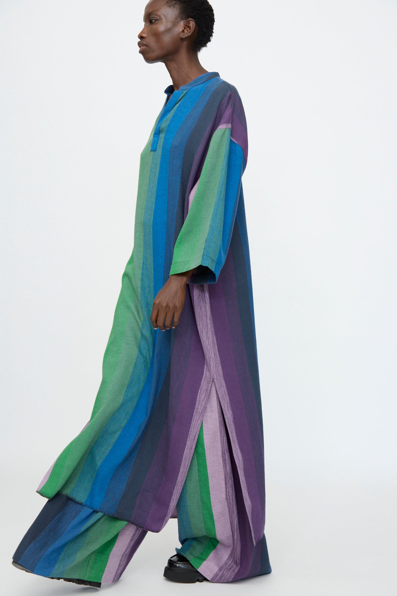 A person wears the Marrakshi Life Nero Collar Caftan in Stripe Multi, an oversized cotton piece in green, blue, and purple stripes, standing against a plain light background.