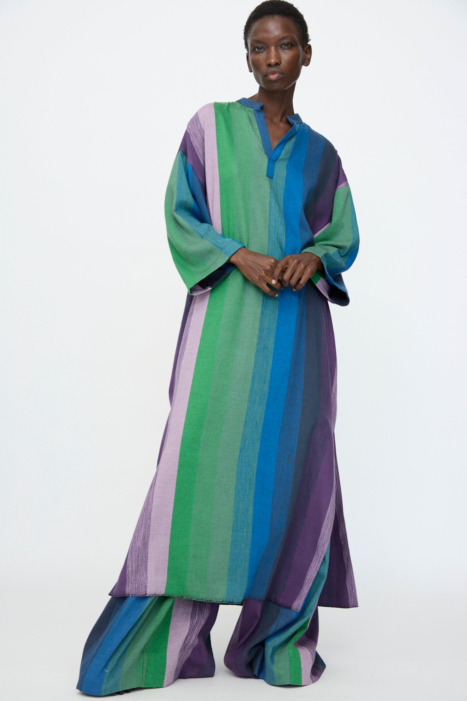 A person wearing the Marrakshi Life Nero Collar Caftan in Stripe Multi, featuring oversized styling with wide vertical stripes in green, blue, and purple, stands against a plain white background.