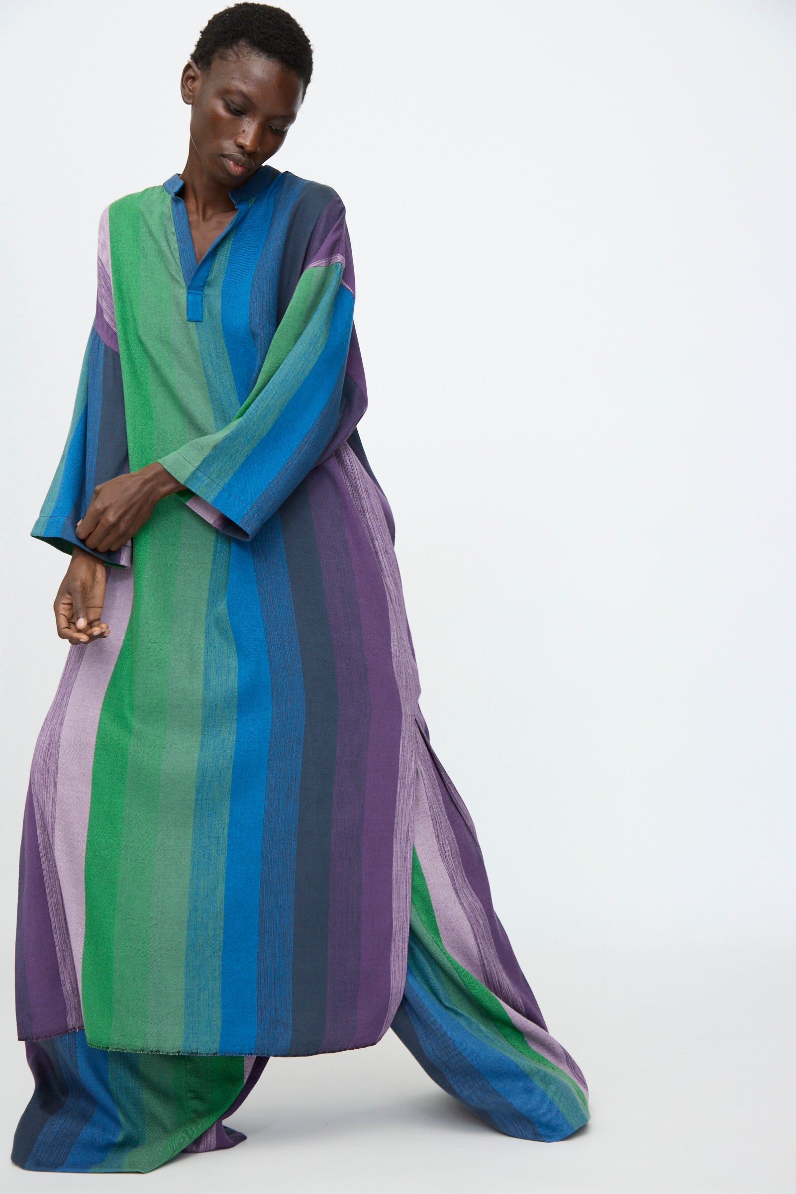 A person wears the Marrakshi Life Nero Collar Caftan in Stripe Multi, featuring oversized vertical blue, green, and purple stripes, standing against a plain light background.