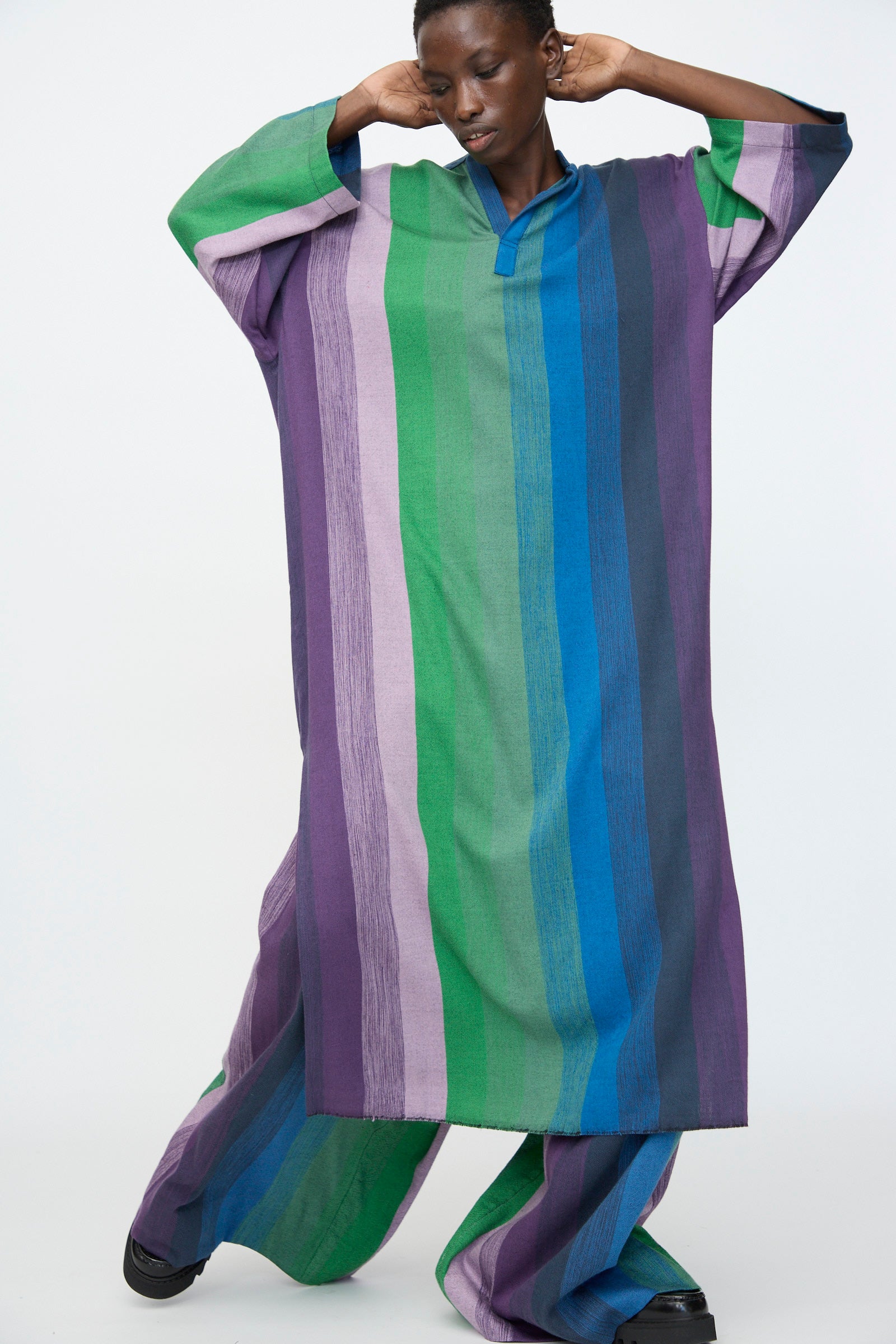 A person wears the Marrakshi Life Nero Collar Caftan in Stripe Multi—featuring loose, long sleeves and wide vertical stripes in green, blue, purple, and lavender—while standing against a plain light background.