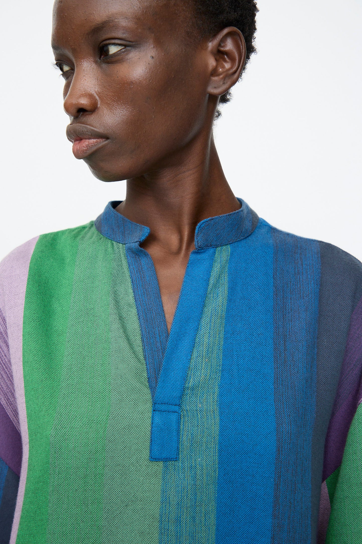 A person wears the Marrakshi Life Nero Collar Caftan in Stripe Multi, featuring vertical multicolored stripes and a short stand-up collar, while looking slightly to the side against a plain background.