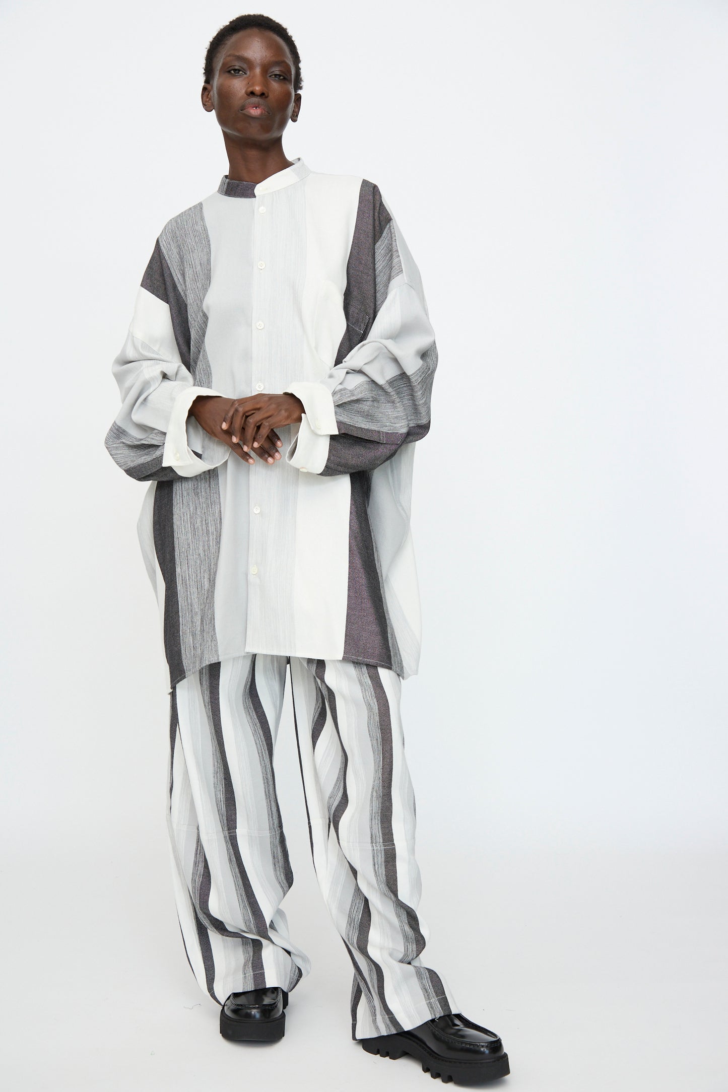 A person wears the Marrakshi Life Nero Collar Oversized Shirt in Grey Stripe—loose-fitting with black, white, and gray vertical stripes—paired with matching wide-leg pants, standing against a plain white background.