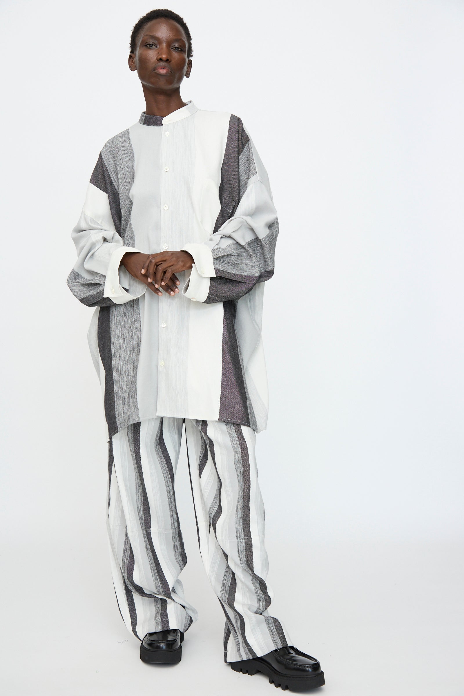A person wears the Marrakshi Life Nero Collar Oversized Shirt in Grey Stripe—loose-fitting with black, white, and gray vertical stripes—paired with matching wide-leg pants, standing against a plain white background.