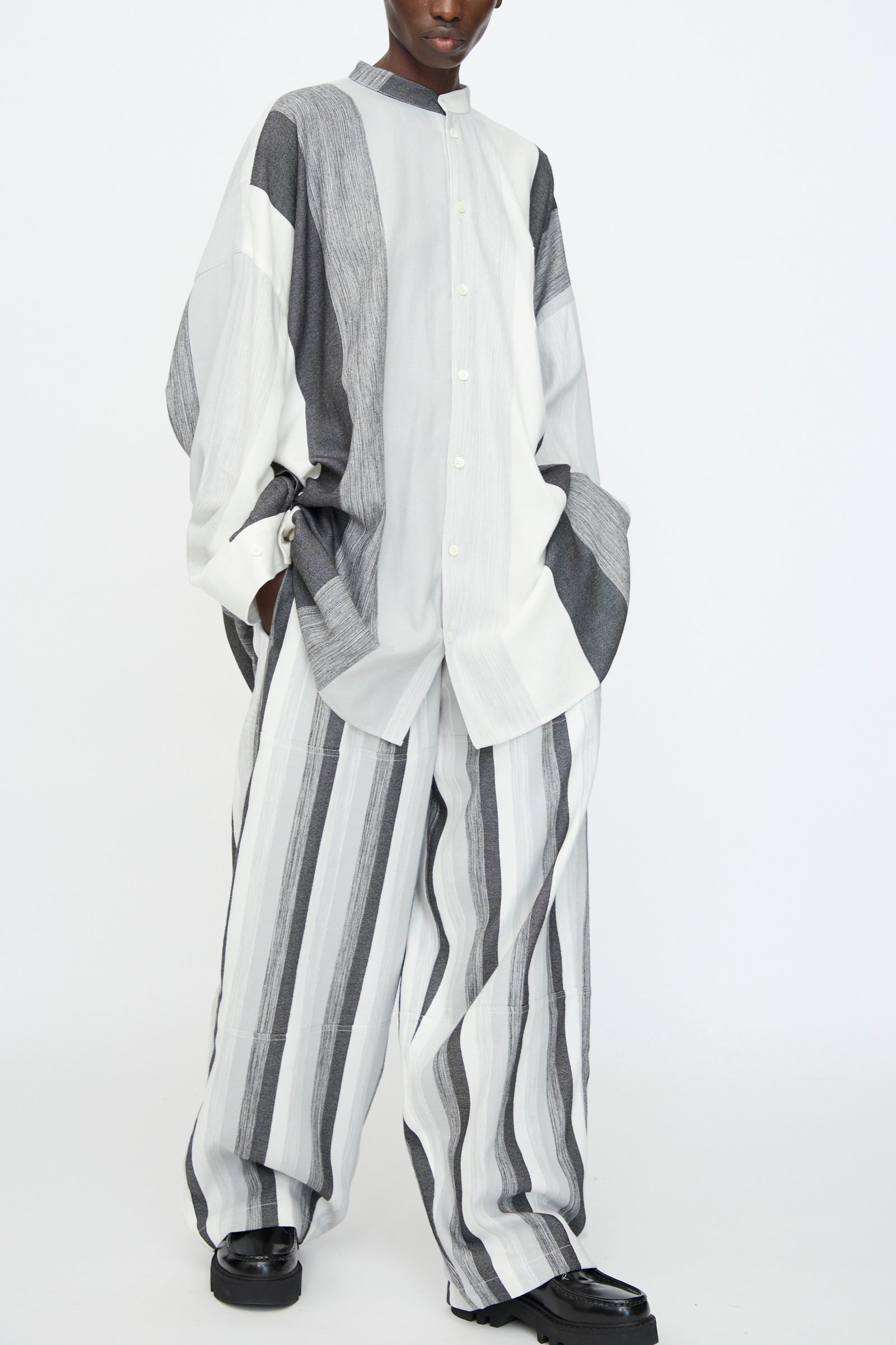 A person wears the Marrakshi Life Nero Collar Oversized Shirt in Grey Stripe, styled with wide-leg striped pants and finished with black shoes.