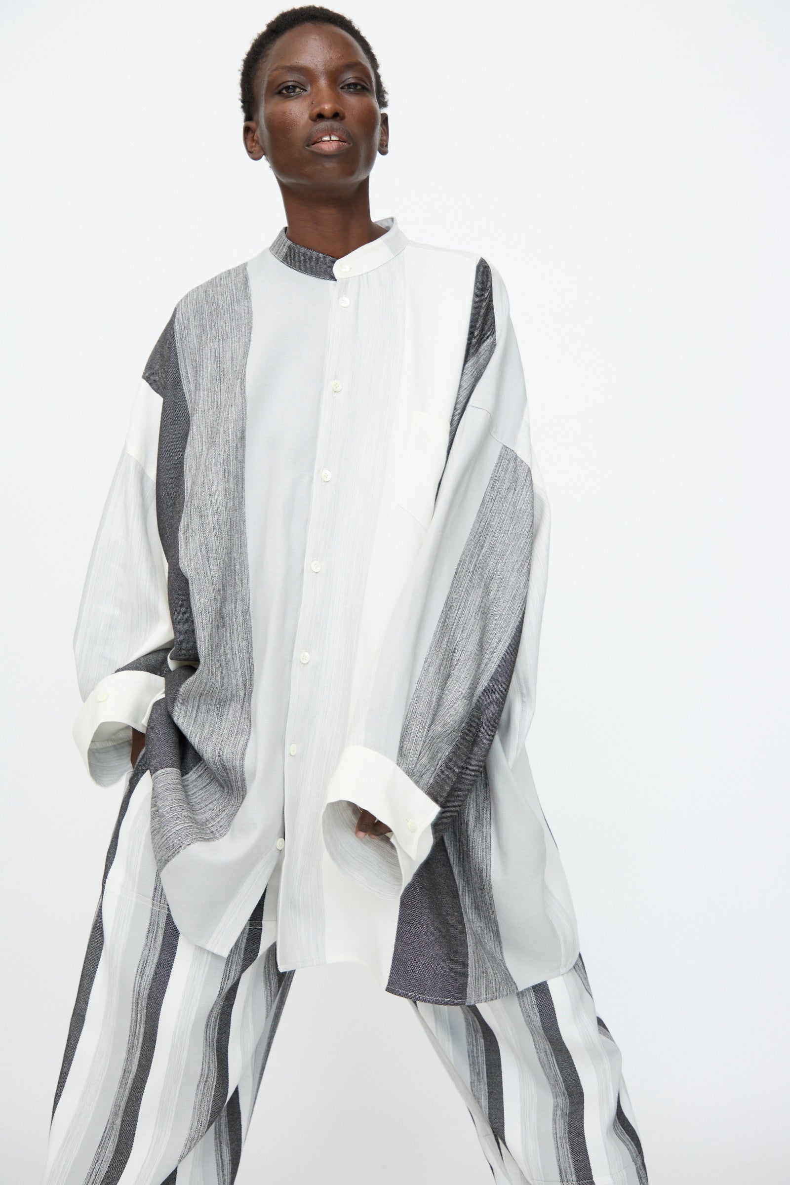 A person wears the Marrakshi Life Nero Collar Oversized Shirt in Grey Stripe—an oversized, long-sleeve cotton shirt with vertical black, white, and gray stripes—paired with matching wide-leg pants against a plain white background.
