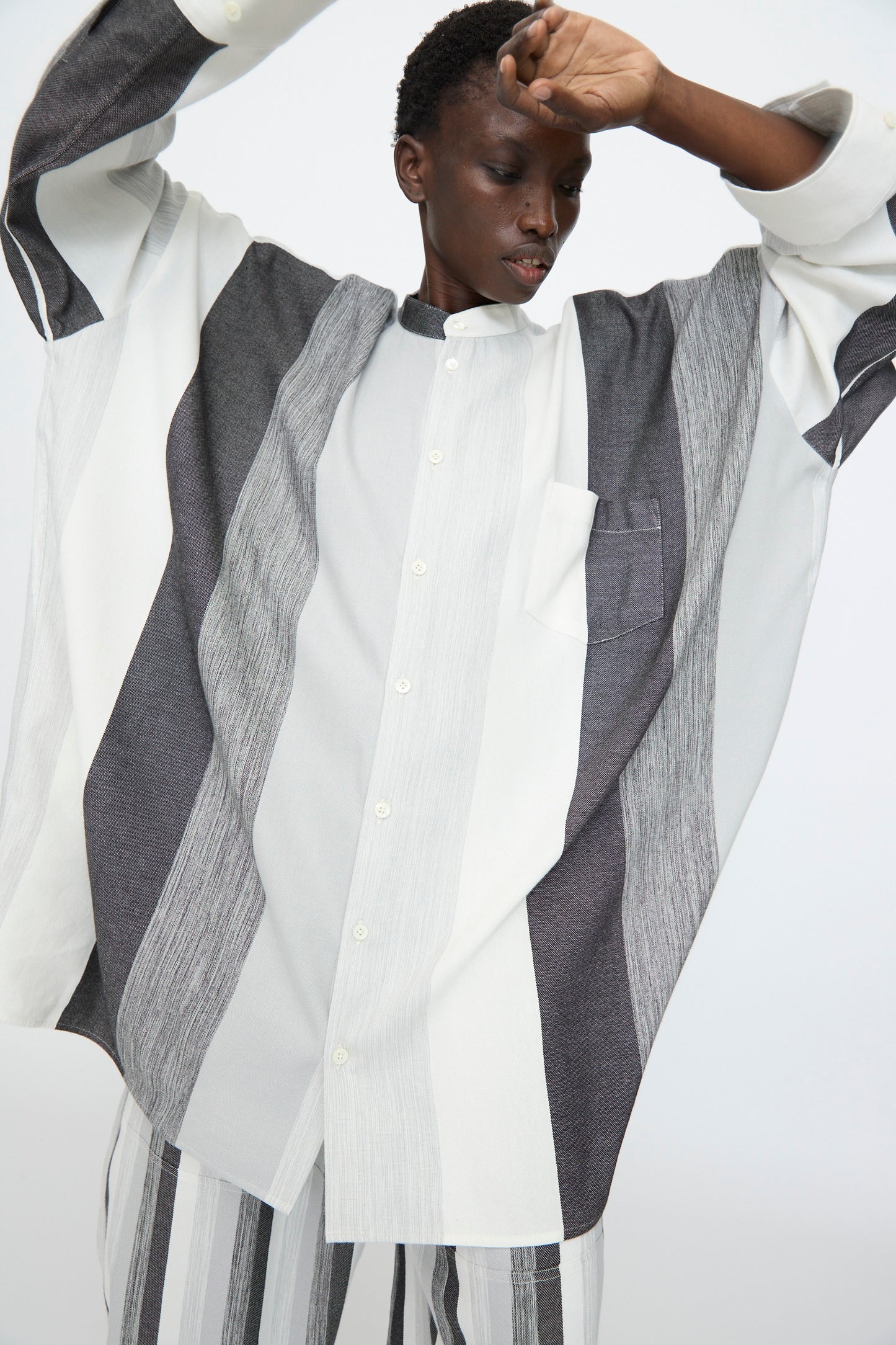 Person wears the Marrakshi Life Nero Collar Oversized Shirt in Grey Stripe with matching pants, featuring a boxy fit and vertical black, gray, and white stripes, standing with one arm raised.
