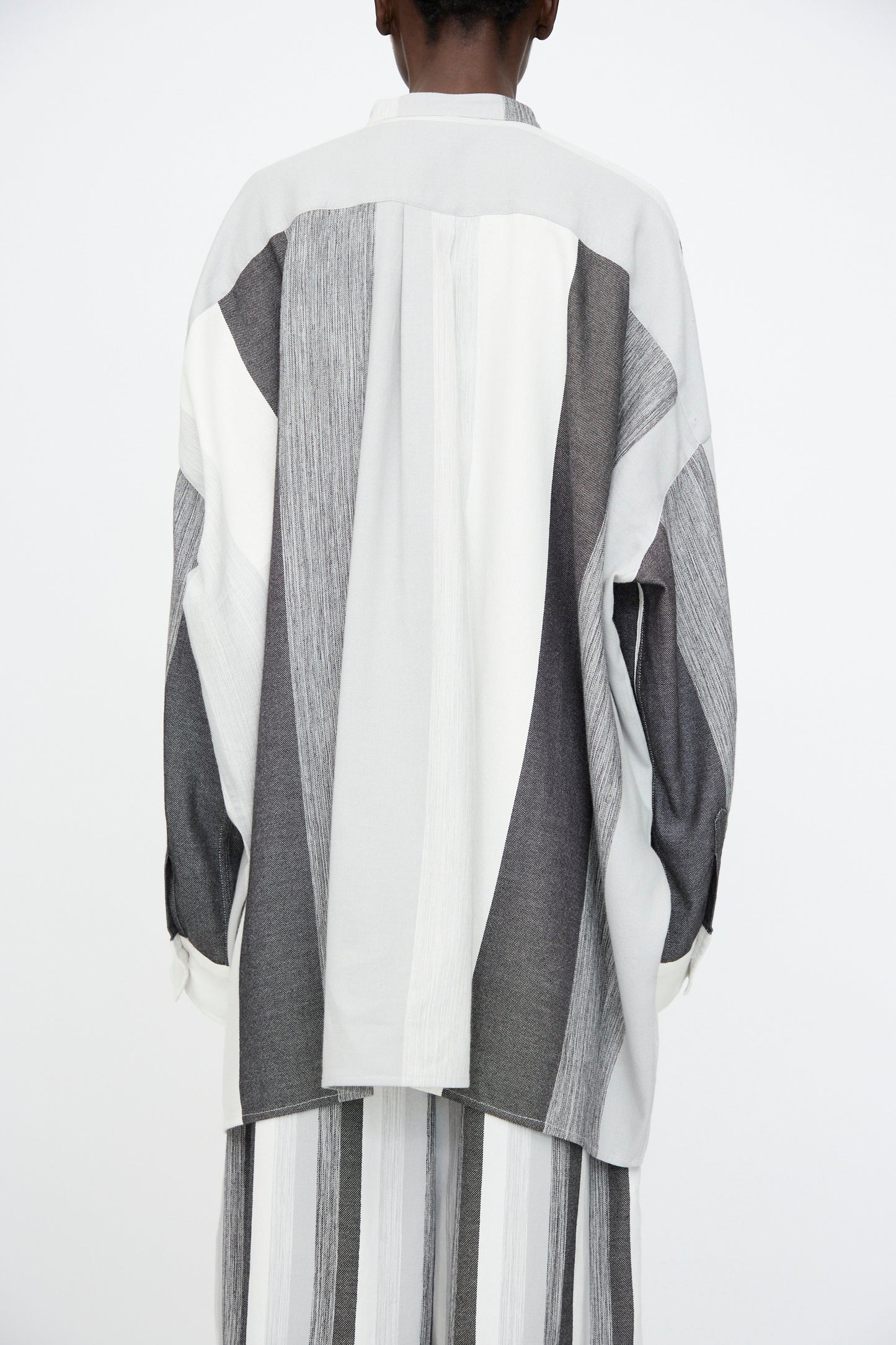 A person wearing the Marrakshi Life Nero Collar Oversized Shirt in Grey Stripe, featuring vertical and diagonal gray and white panels, paired with matching striped pants, shown from the back against a plain background.