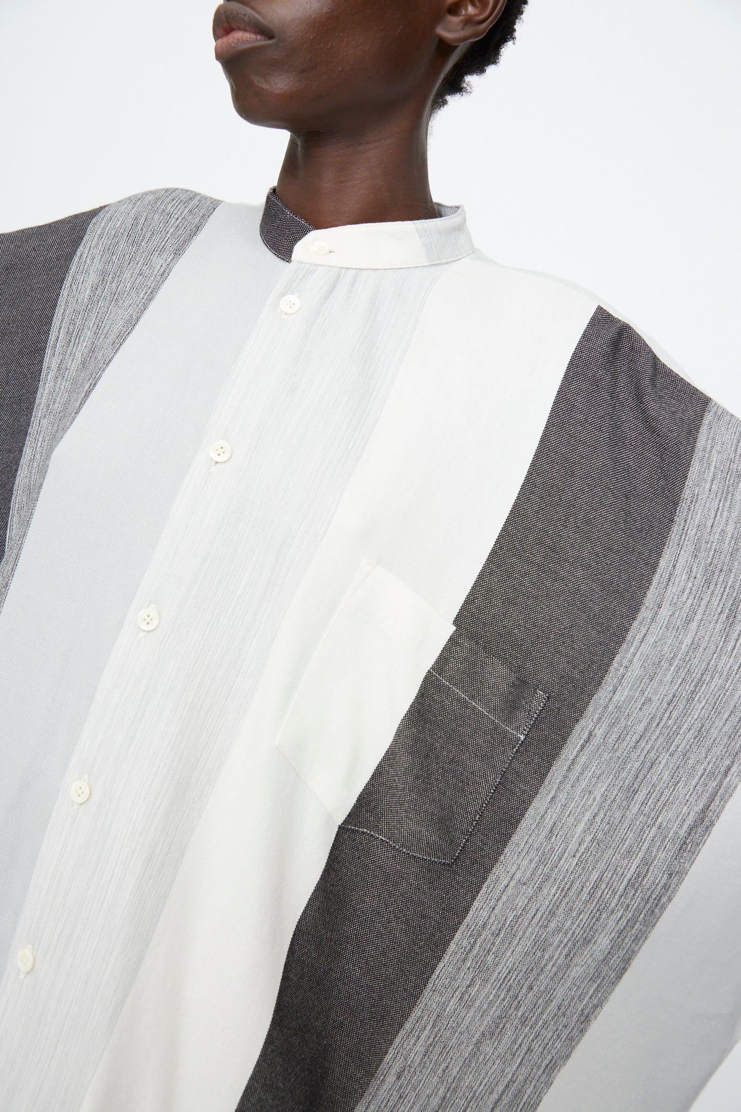 A person wearing the Marrakshi Life Nero Collar Oversized Shirt in Grey Stripe, featuring wide vertical panels in white, grey, and charcoal, a stand collar, and a chest pocket.