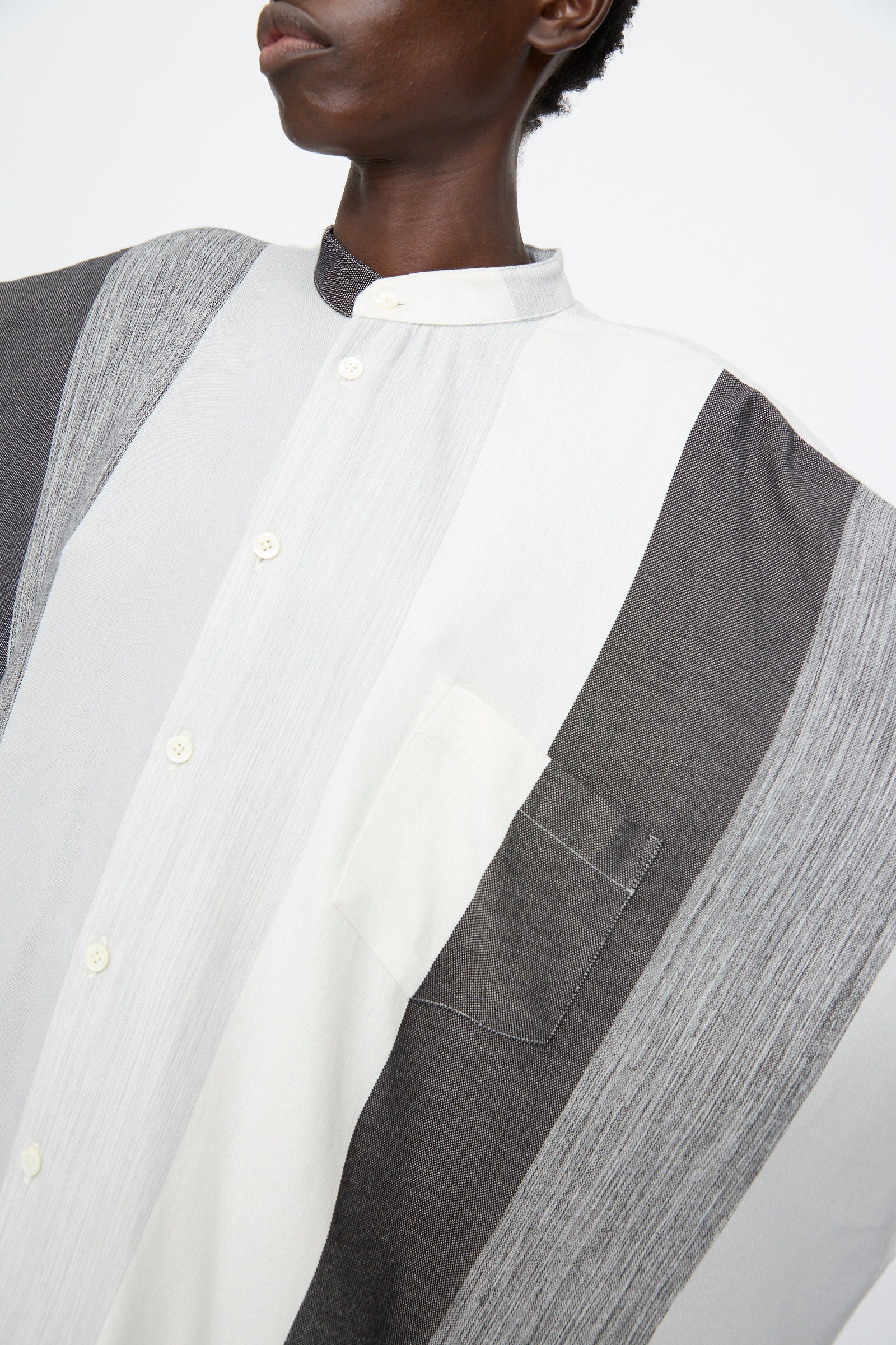 A person wearing the Marrakshi Life Nero Collar Oversized Shirt in Grey Stripe, featuring wide vertical panels in white, grey, and charcoal, a stand collar, and a chest pocket.