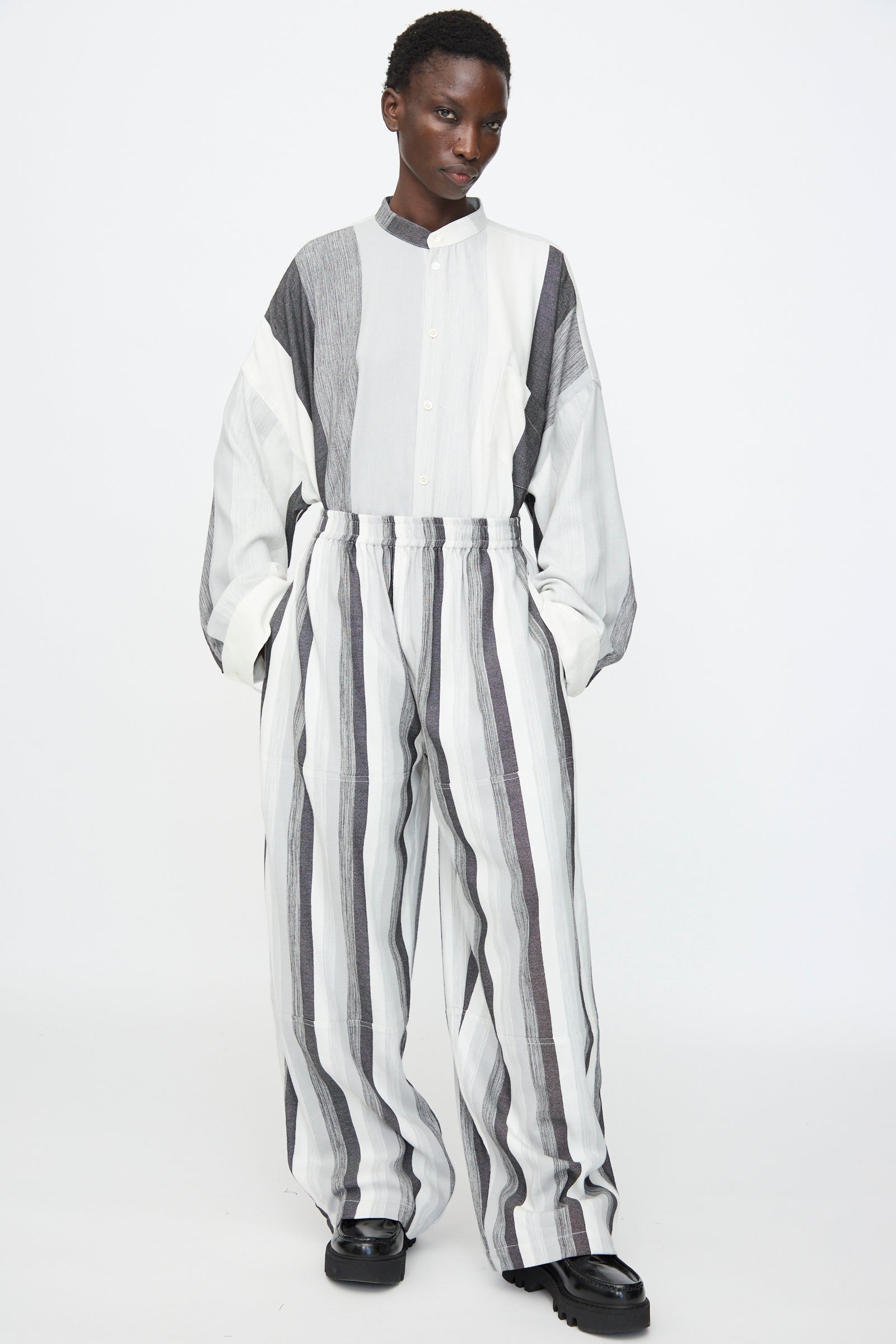 A person wears Marrakshi Life's Parachute Pant in Grey Stripe with an oversized, striped black, white, and gray button-up shirt, black shoes, and stands against a plain background.