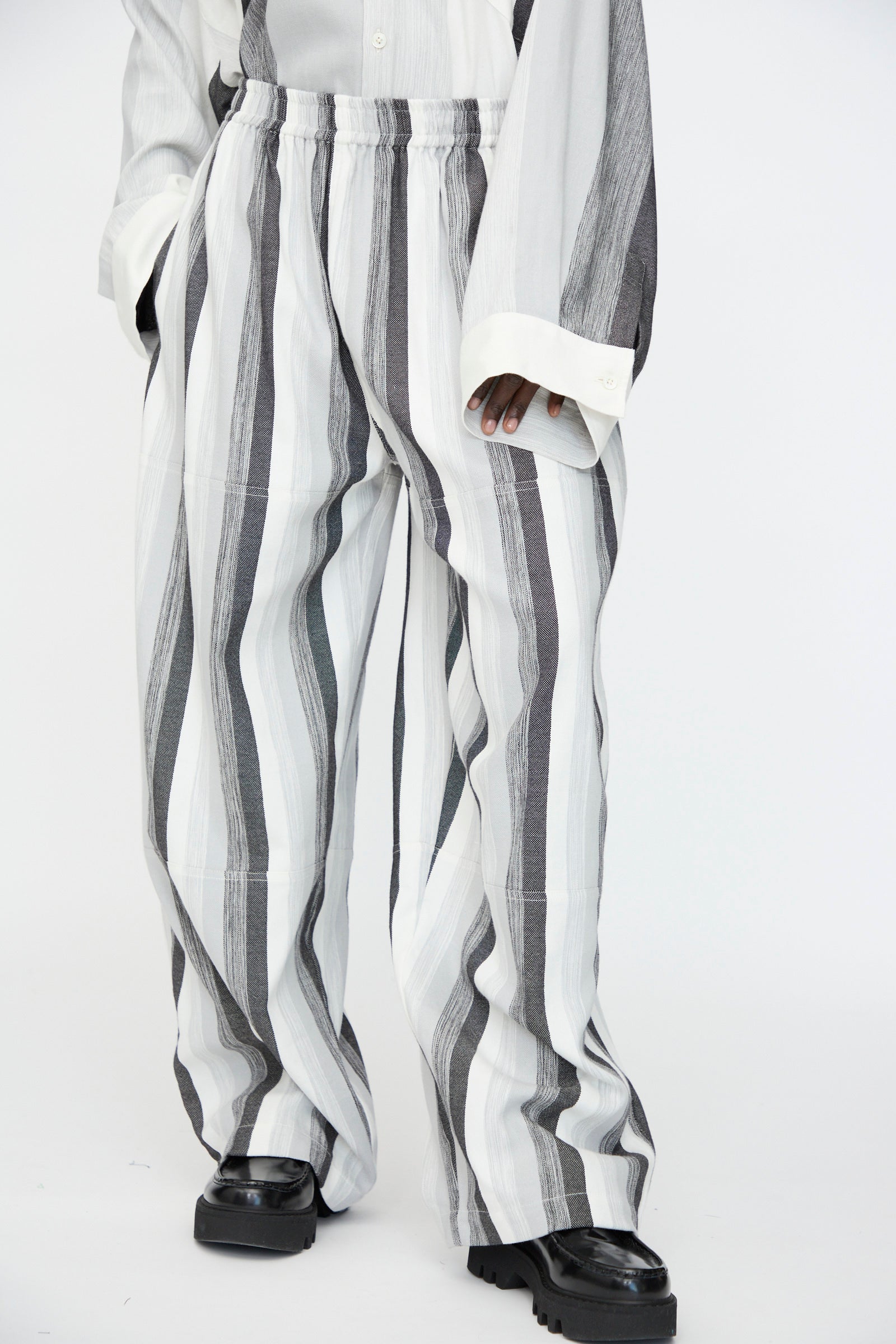 Person wearing Marrakshi Life's Parachute Pant in Grey Stripe—an oversized, wide-leg cotton pant with vertical black and white stripes—paired with a matching shirt and chunky black shoes, shown from shoulders down.