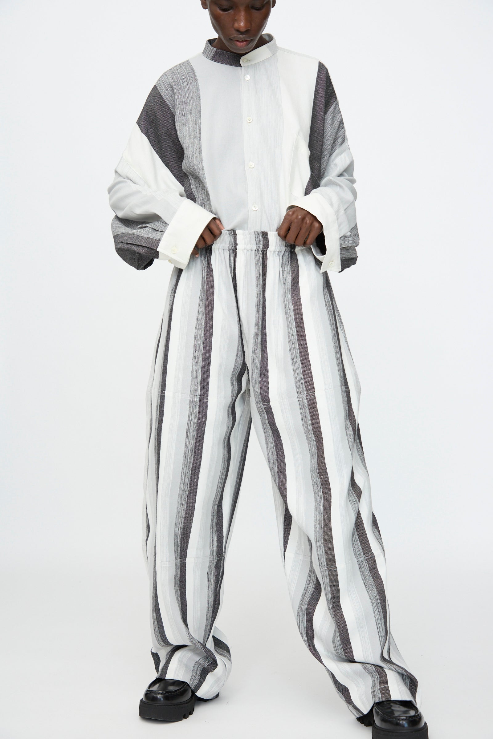 A person wears the Marrakshi Life Parachute Pant in Grey Stripe—wide-leg with vertical gray and white stripes—paired with a striped cotton oversized button-up shirt, standing against a plain white background.