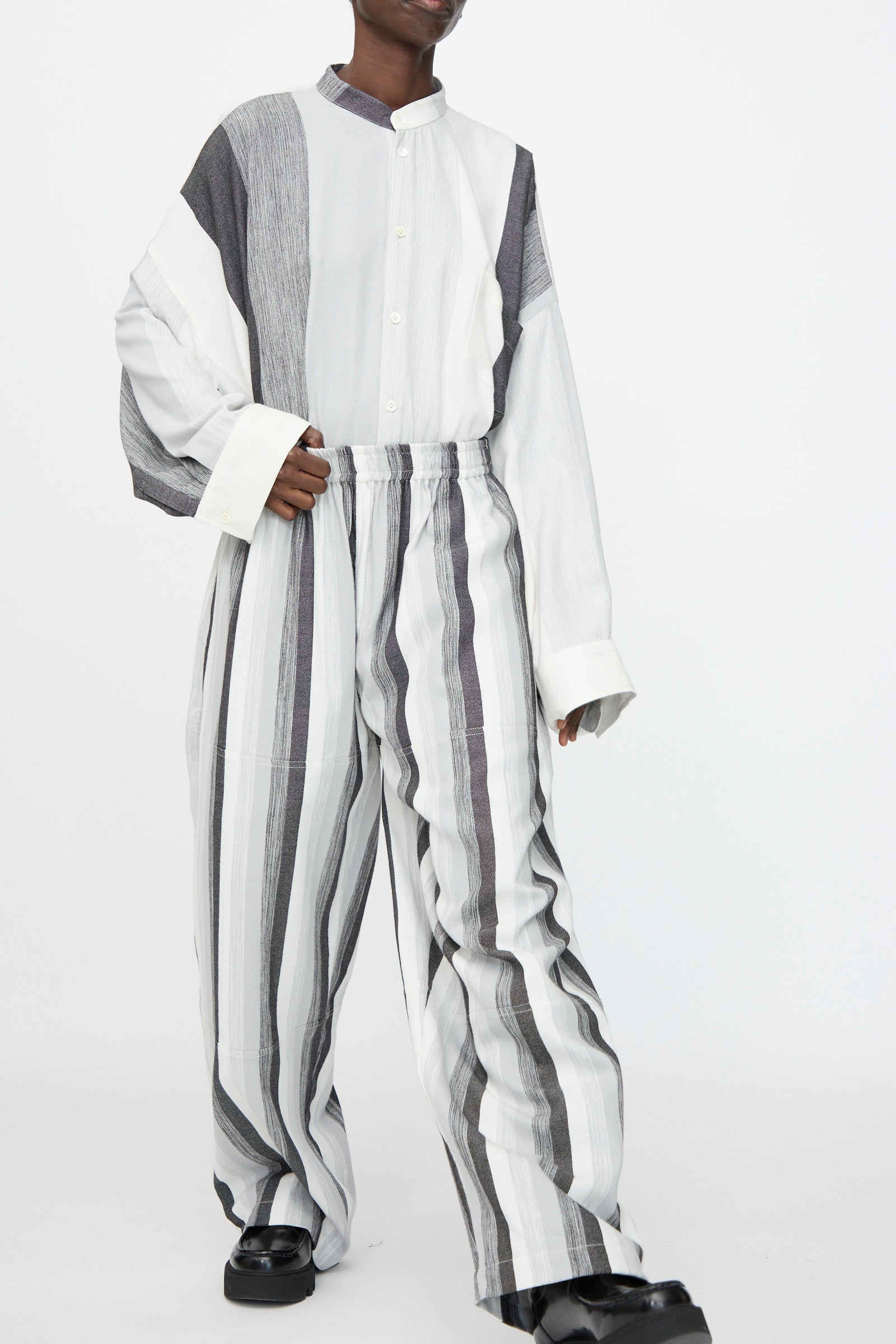 Person stands against a white background, wearing Marrakshi Life's Parachute Pant in Grey Stripe—an oversized fit, wide-leg style with vertical black, gray, and white stripes—paired with a matching long-sleeve button-up shirt.