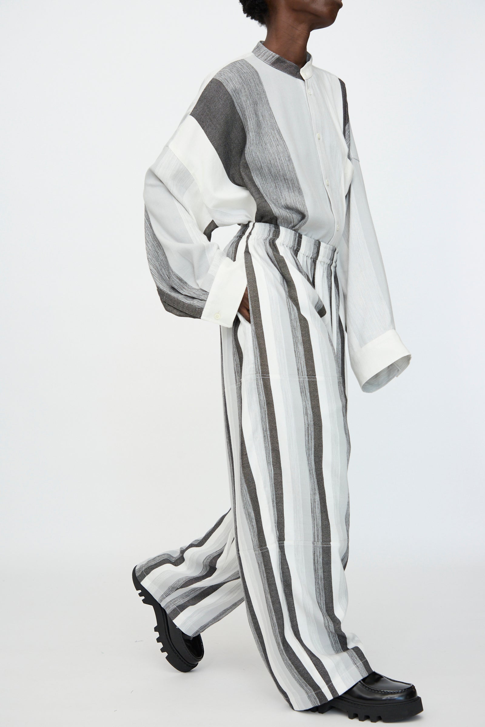 Model wears Marrakshi Life's Parachute Pant in Grey Stripe—an oversized, wide leg style—paired with a long-sleeved, button-up striped cotton shirt, walking against a plain white background.
