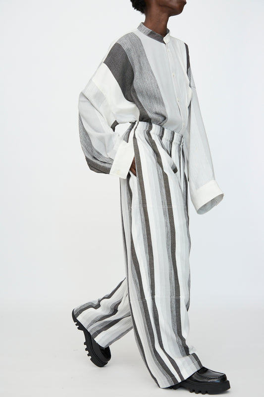 Model wears Marrakshi Life's Parachute Pant in Grey Stripe—an oversized, wide leg style—paired with a long-sleeved, button-up striped cotton shirt, walking against a plain white background.