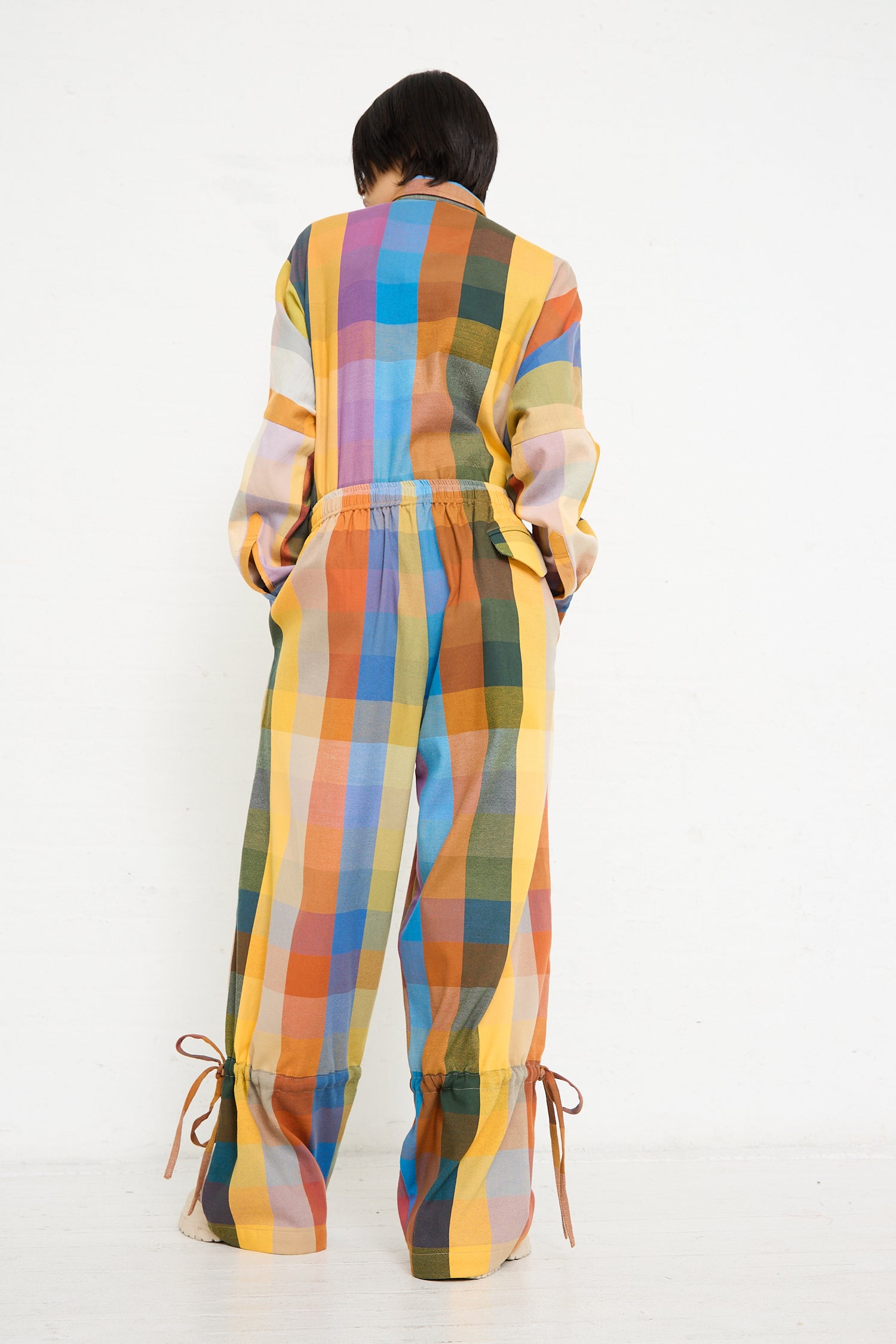 A person standing with their back to the camera, wearing a colorful striped outfit designed by Marrakshi Life with an elastic waist and tie details at the ankles, highlighting an oversized Parachute Pant in Multi Check fit.