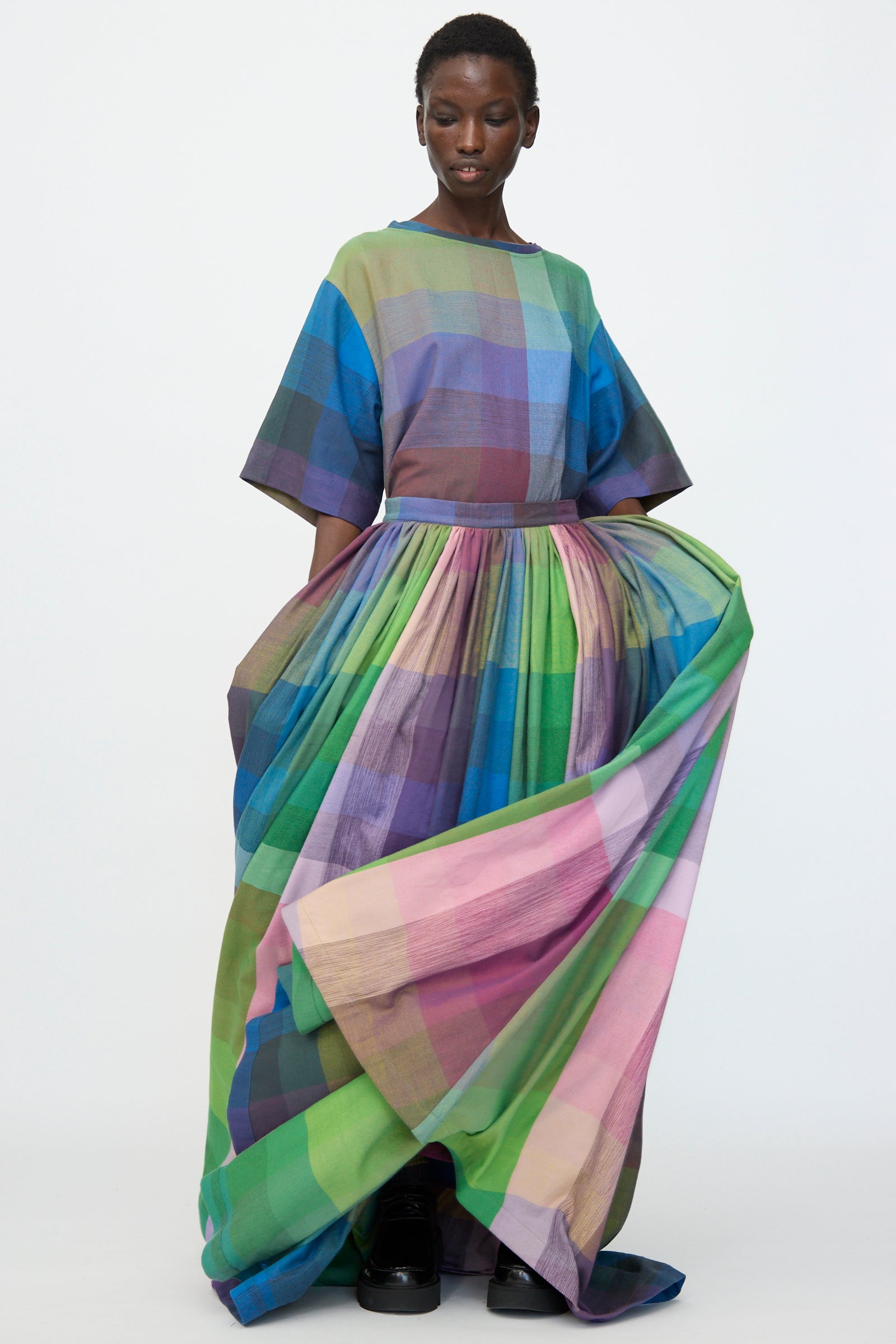 A person wears the Marrakshi Life Puffer Skirt in Plaid Multi, a floor-length cotton design with wide sleeves and a vibrant blue, green, purple, and pink pattern, standing against a plain white background.