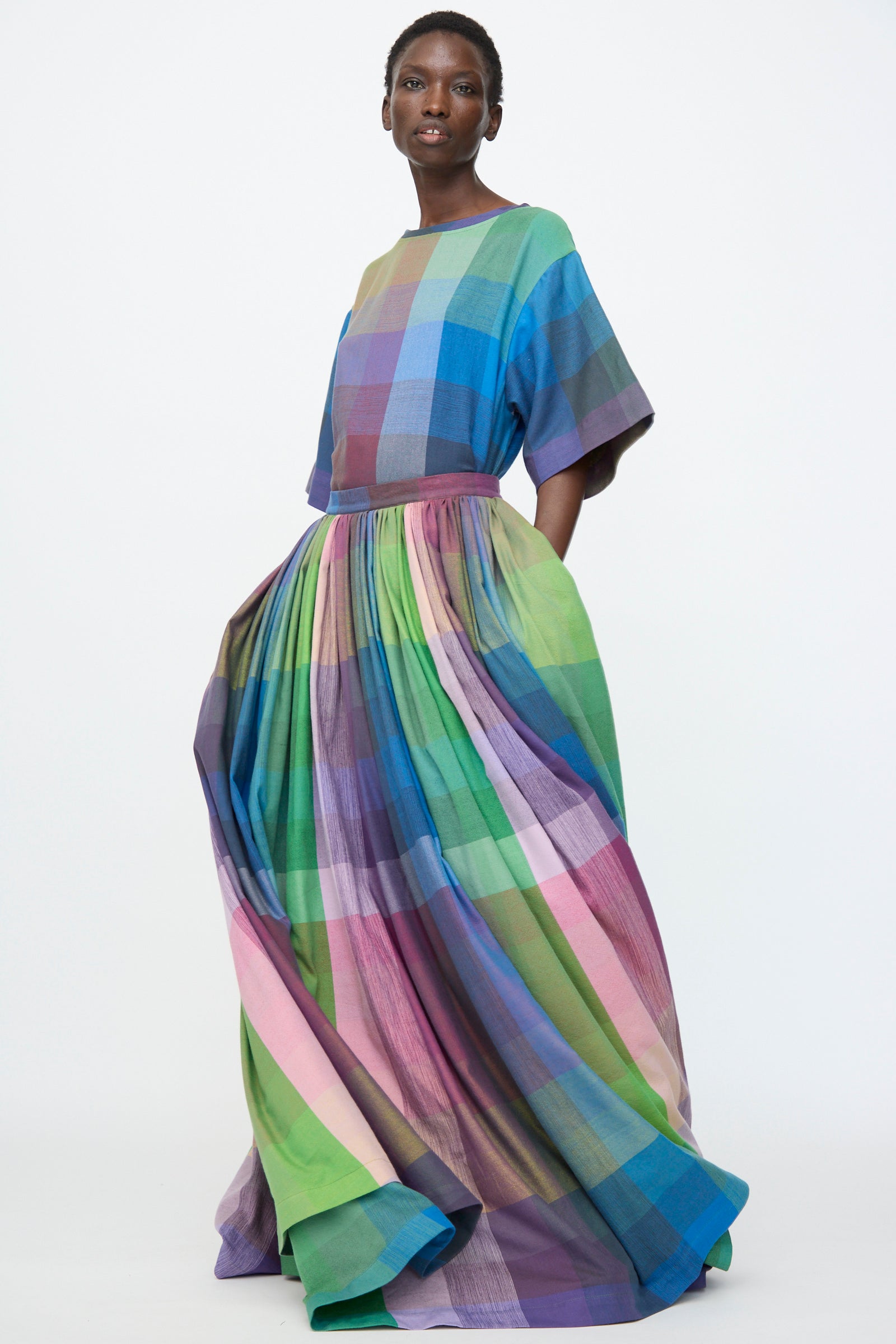 A person wears the Marrakshi Life Puffer Skirt in Plaid Multi, a floor-length plaid cotton skirt with short sleeves and a full skirt, standing against a plain white background.