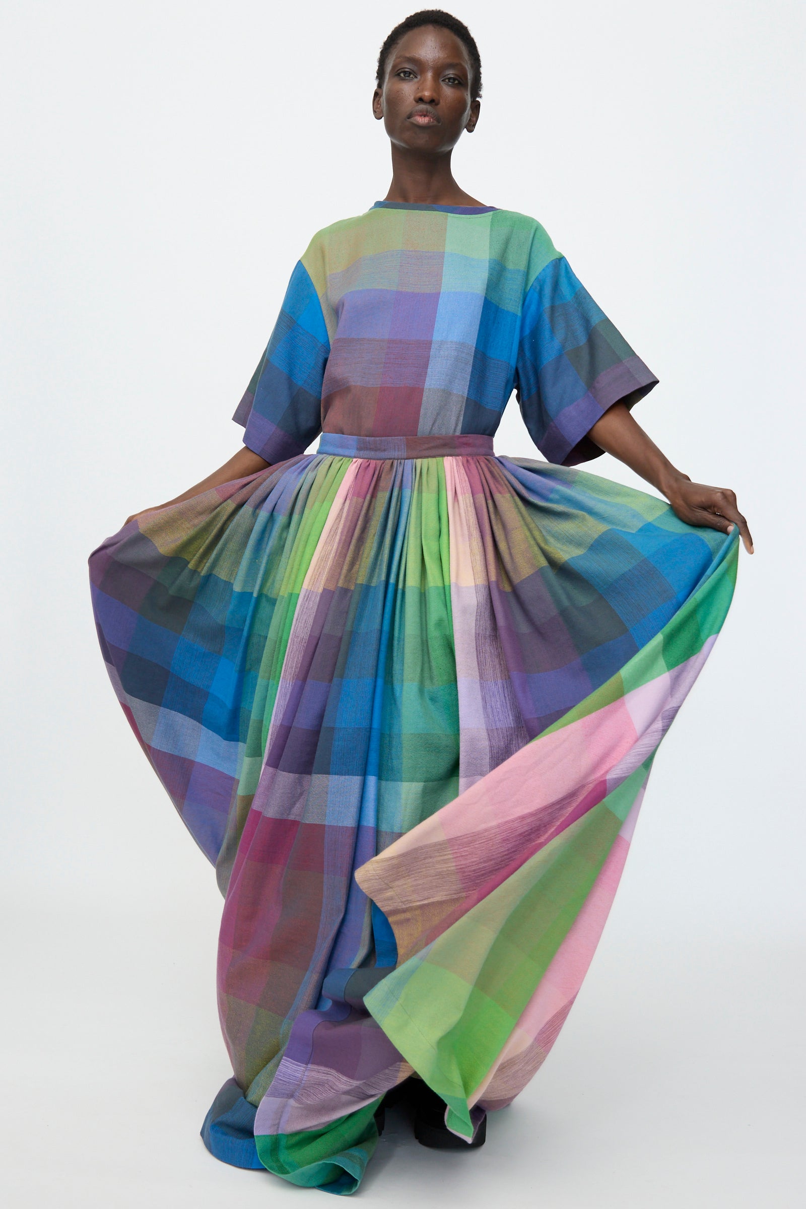 A person stands wearing the Marrakshi Life Puffer Skirt in Plaid Multi, a floor-length cotton maxi with bold multicolored plaid, holding out the skirt's sides.