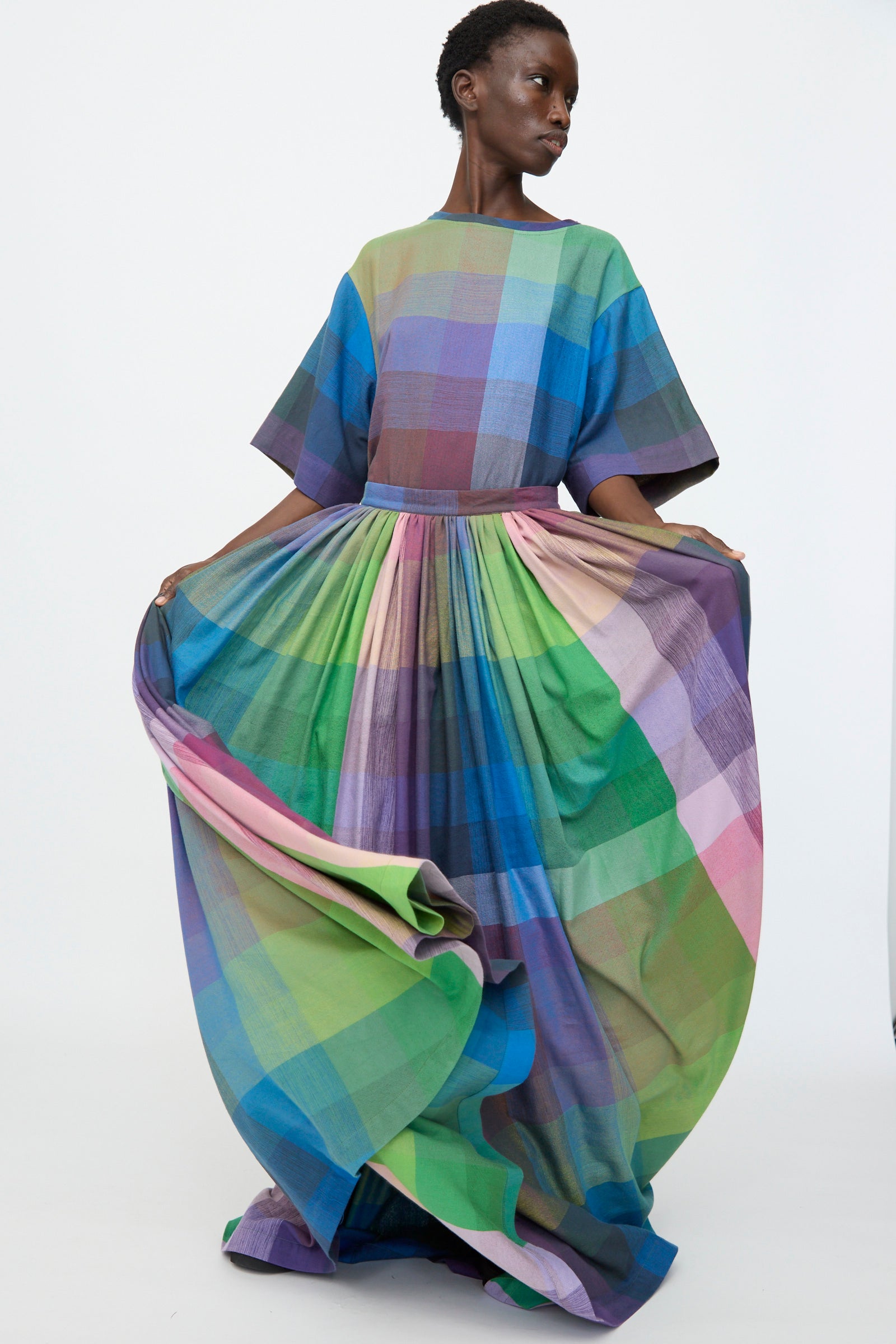 A person wearing the Marrakshi Life Puffer Skirt in Plaid Multi holds out the colorful cotton skirt, showcasing its floor-length design and vibrant green, blue, purple, and pink plaid pattern against a plain background.