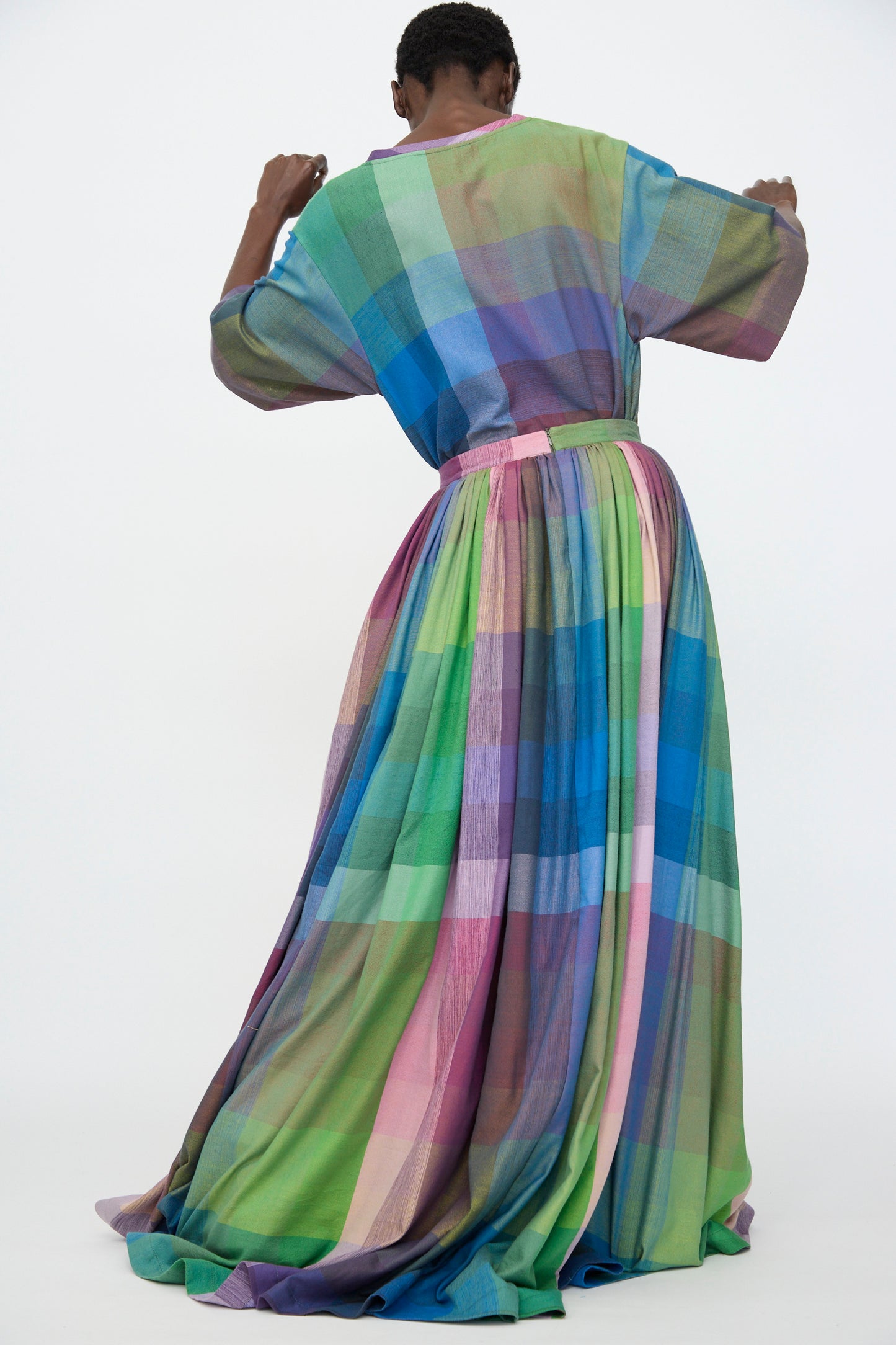A person is photographed from behind in Marrakshi Life’s Puffer Skirt in Plaid Multi—a long, colorful plaid skirt—against a plain white background.