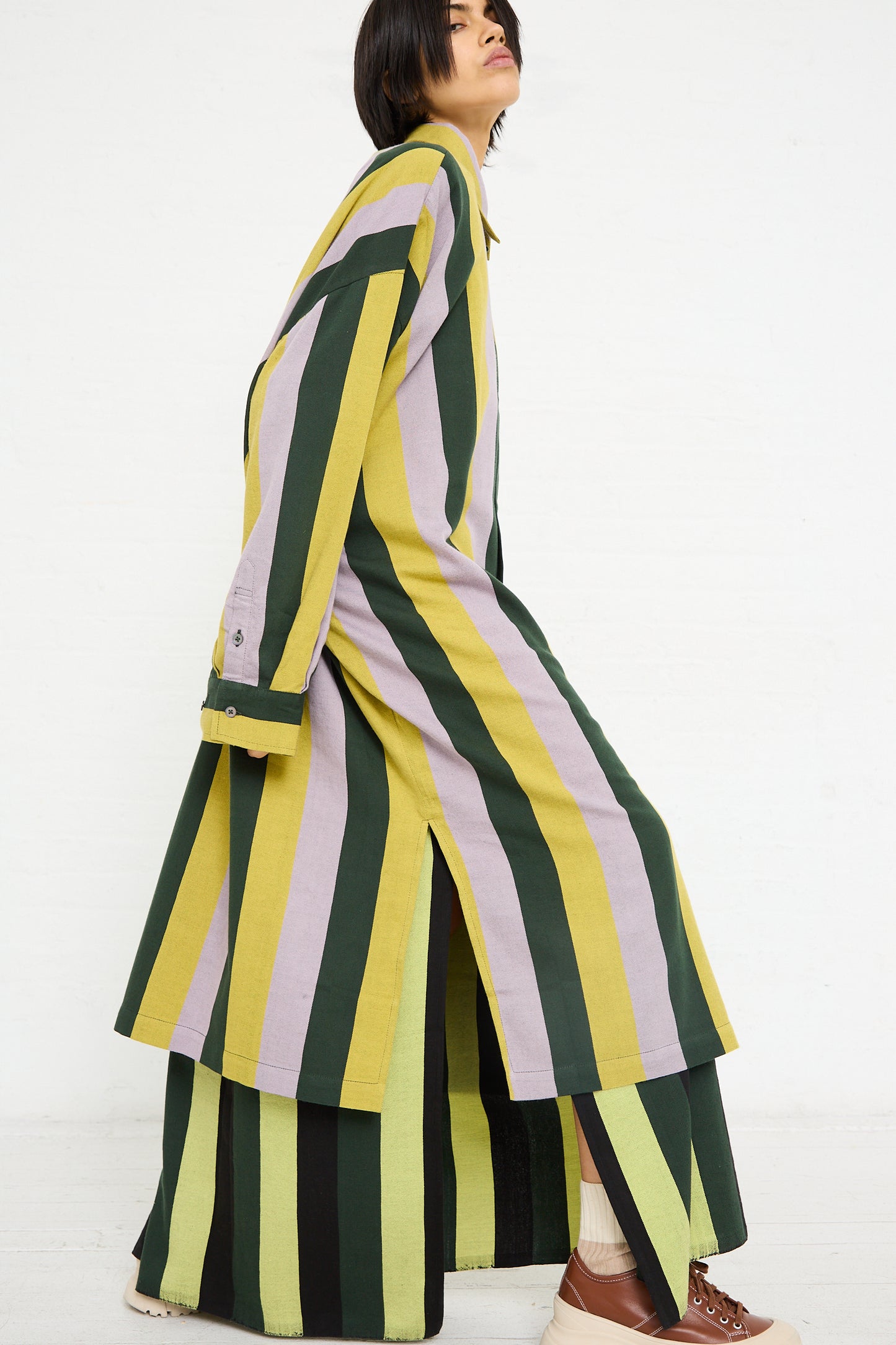 A woman in a Marrakshi Life Small Collar Oversized Shirt in Stripe, embracing traditional Moroccan wardrobe aesthetics with green, yellow, and black stripes, standing in profile against a white background.