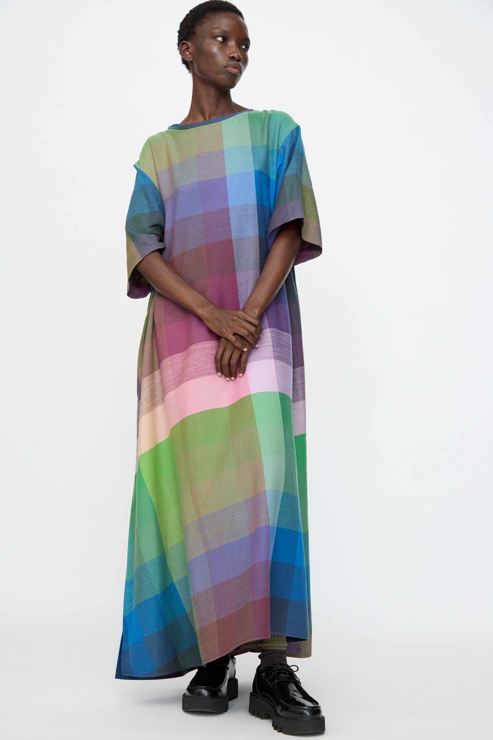 A person wearing the Marrakshi Life T Shirt Dress in Plaid Multi, styled in an oversized ankle-length fit with short sleeves and black platform shoes, stands against a plain white background.