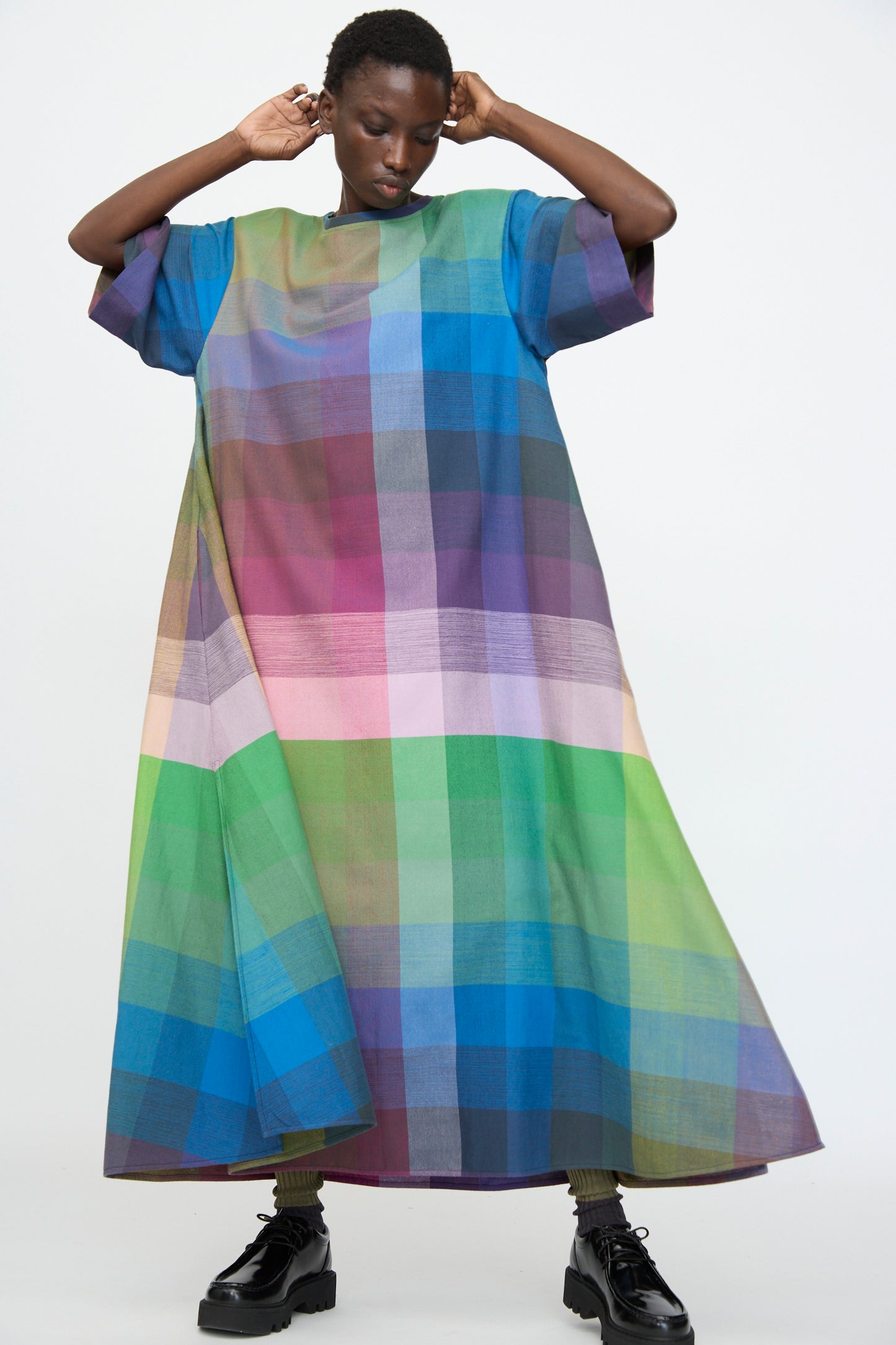 A person wearing the Marrakshi Life T Shirt Dress in Plaid Multi, an oversized, floor-length cotton dress with a colorful blue, green, purple, and pink plaid pattern, stands against a plain white background.