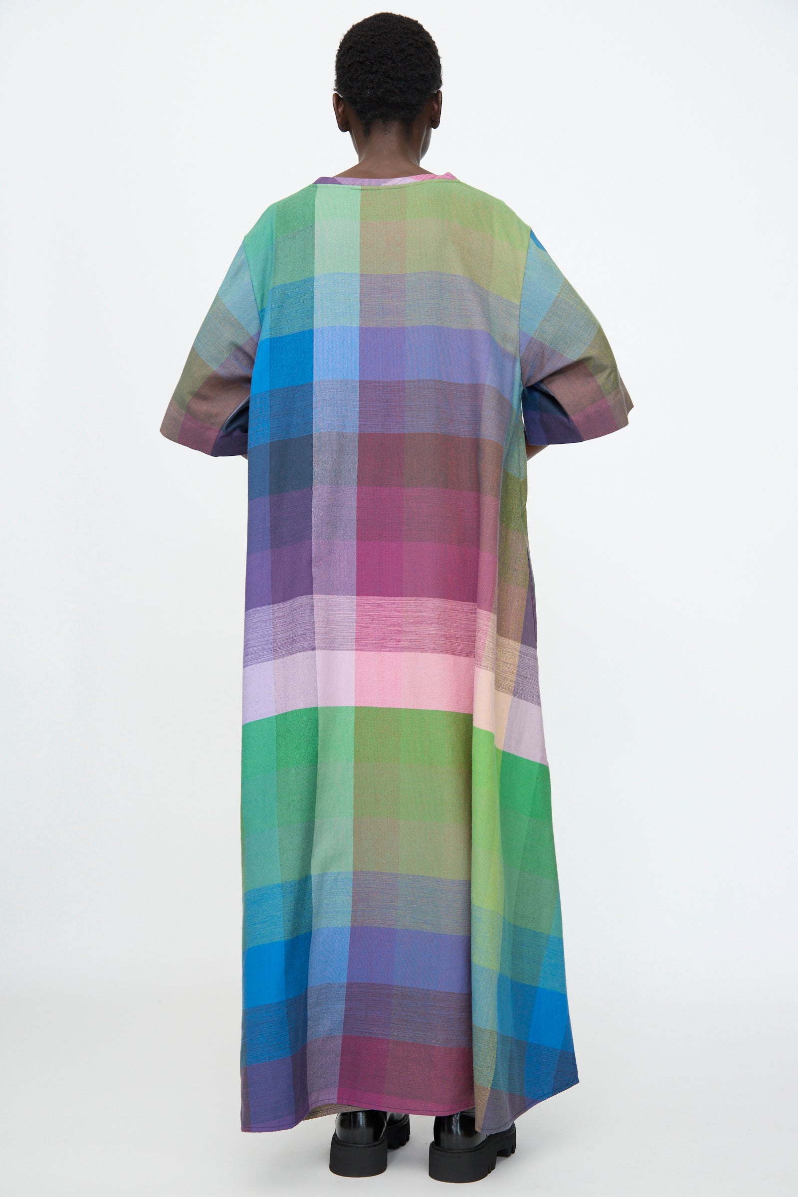 A person stands with their back to the camera, wearing the Marrakshi Life T Shirt Dress in Plaid Multi—an oversized, loose cotton dress featuring a blend of green, blue, purple, pink, and red shades.