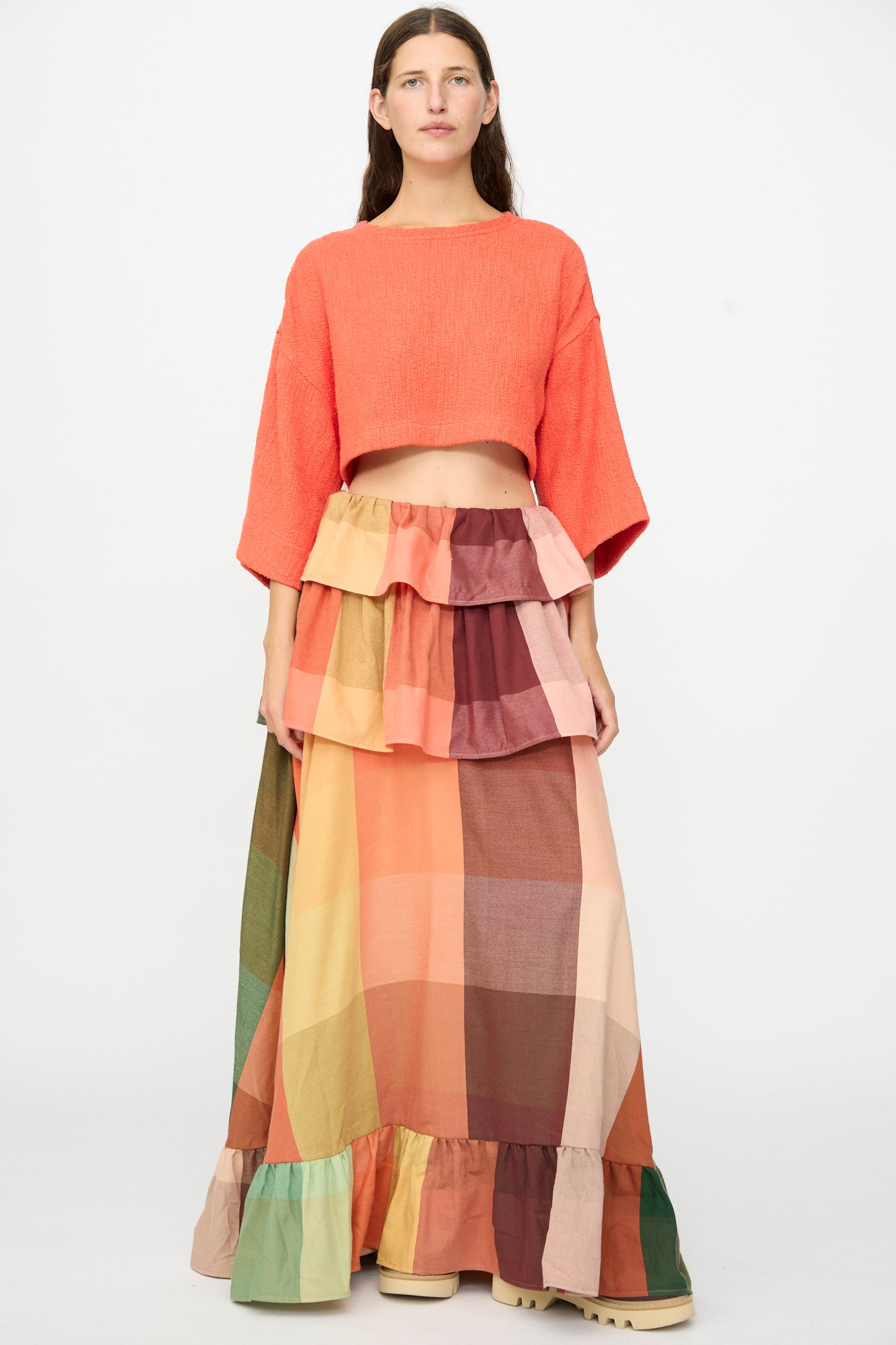 A woman stands against a white background wearing Marrakshi Life’s The Cropped T-Shirt in Burnt Orange Flamme, styled with a long, multi-colored plaid tiered skirt featuring ruffle details and crafted from handwoven cotton.