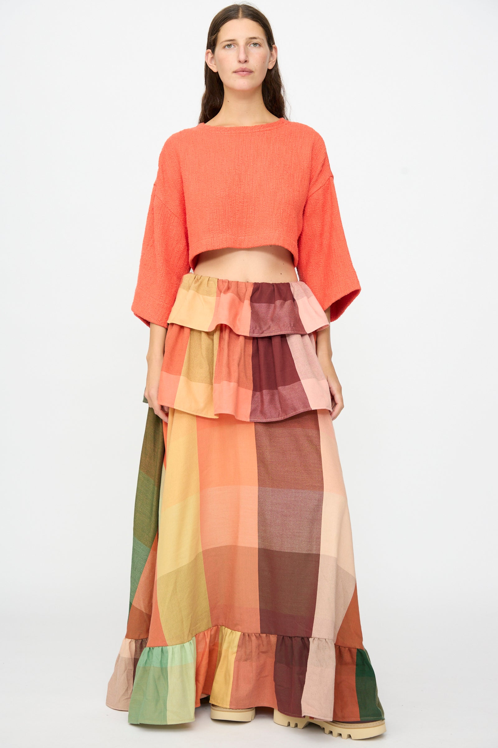 A woman stands against a white background wearing Marrakshi Life’s The Cropped T-Shirt in Burnt Orange Flamme, styled with a long, multi-colored plaid tiered skirt featuring ruffle details and crafted from handwoven cotton.