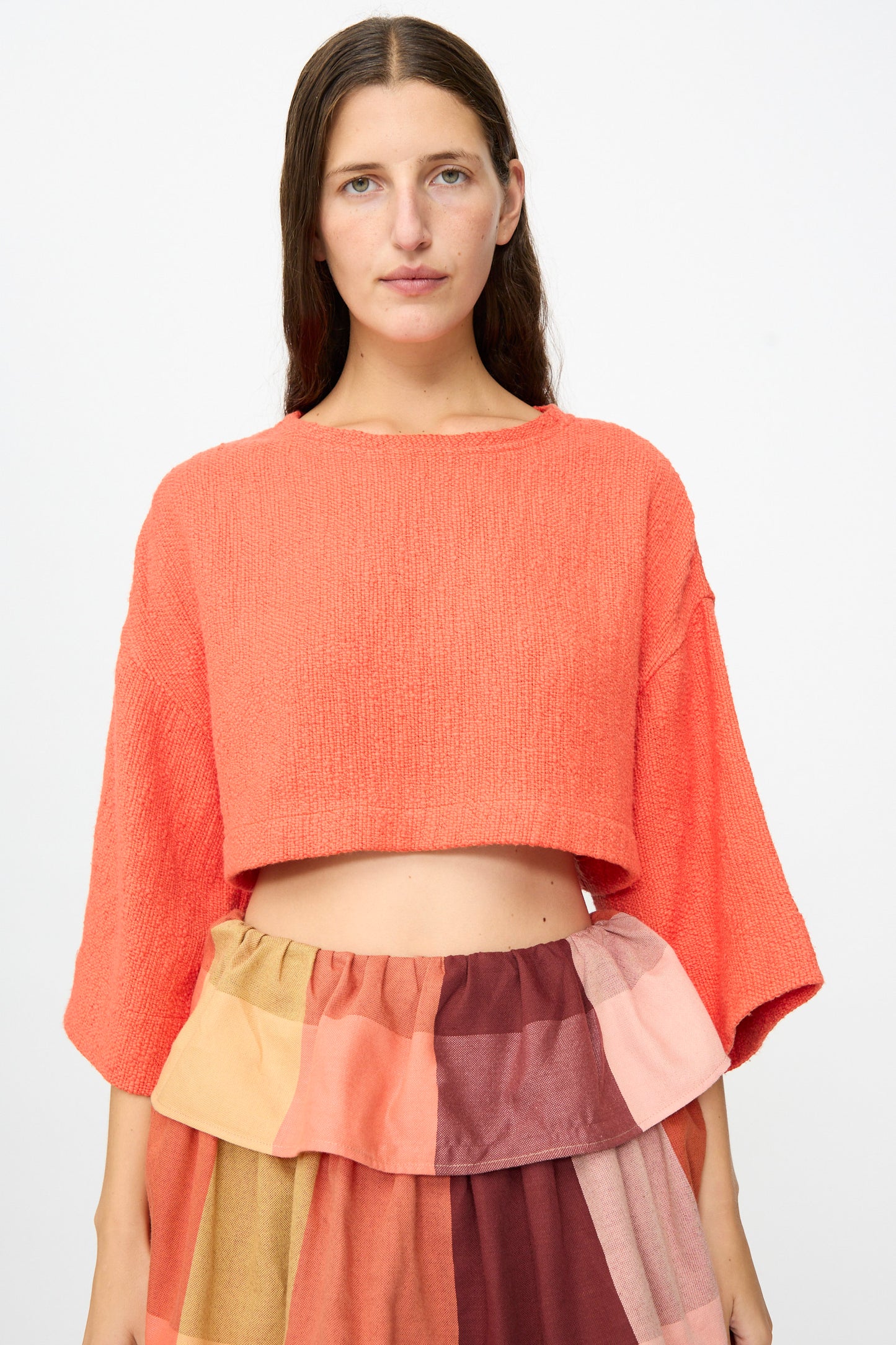 A woman wearing Marrakshi Life’s Cropped T-Shirt in Burnt Orange Flamme and a multicolored patchwork skirt made from handwoven cotton, standing against a plain white background.