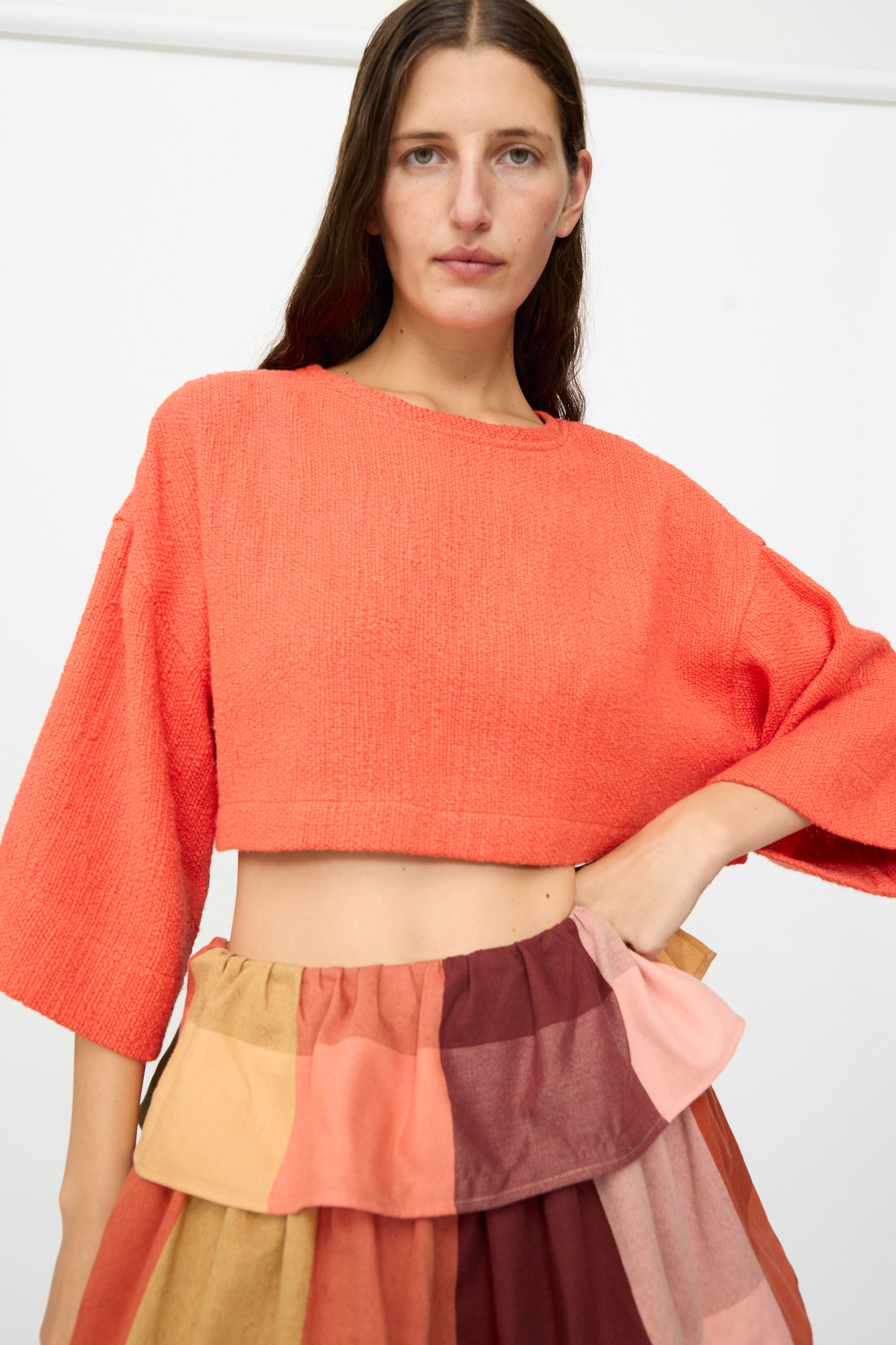 A woman wears Marrakshi Life’s Cropped T-Shirt in Burnt Orange Flamme and a multi-colored patchwork skirt, posing against a plain white background.
